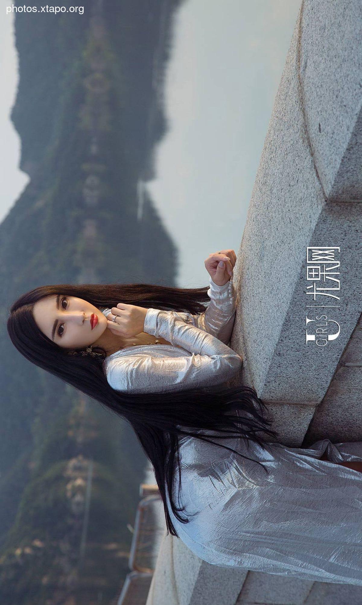 A woman in a silver dress sits on a stone wall overlooking a mountain range.  Her long dark hair cascades down her back. The image has a moody and atmospheric quality.