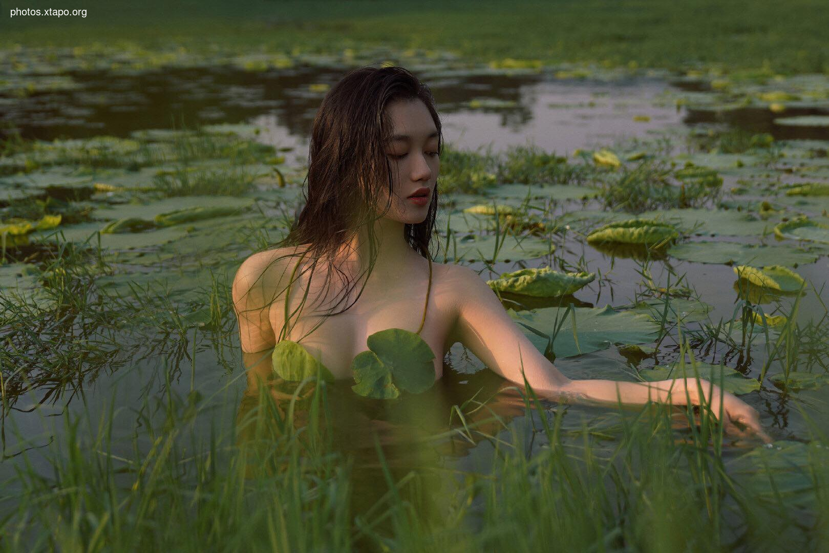 A woman with long dark hair emerges from a serene pond covered in lily pads. The soft light and lush greenery create a mystical atmosphere.
