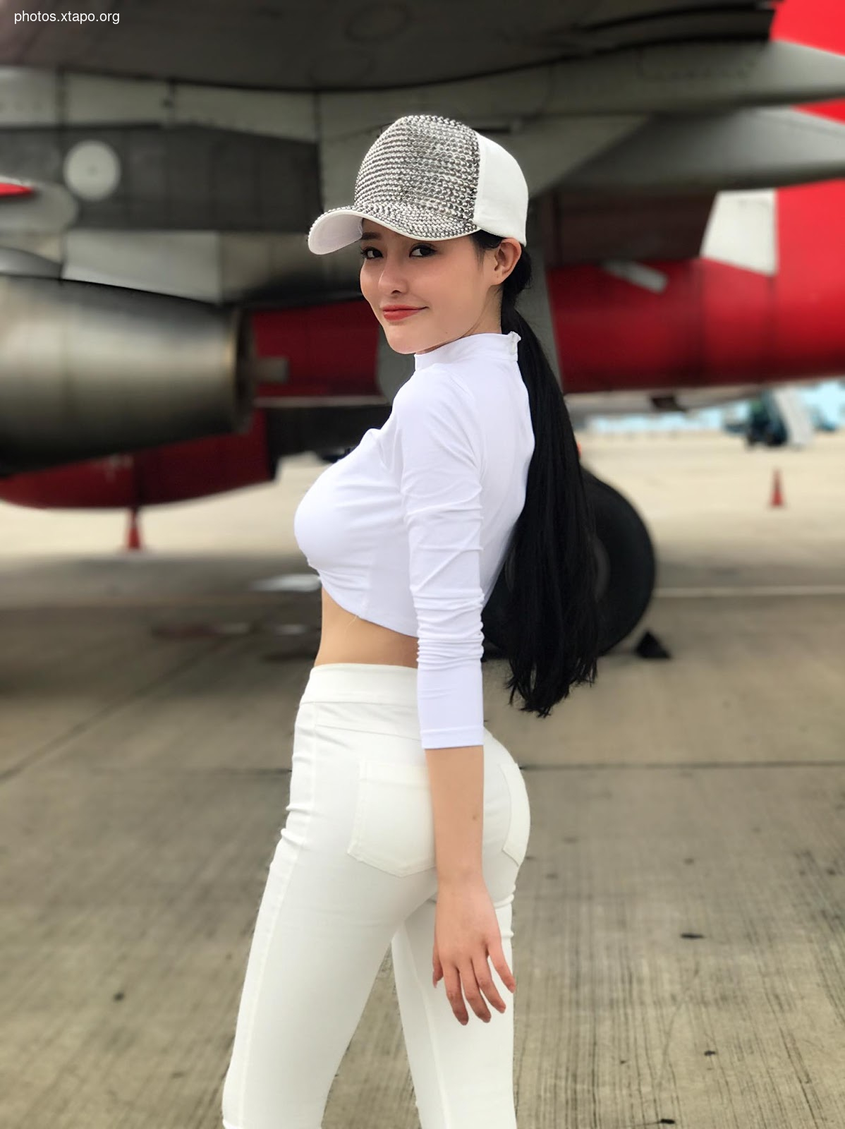 A captivating young woman, elegantly dressed in a white crop top and pants, poses confidently near a red airplane. Her stylish rhinestone-studded baseball cap adds a touch of flair to her look.