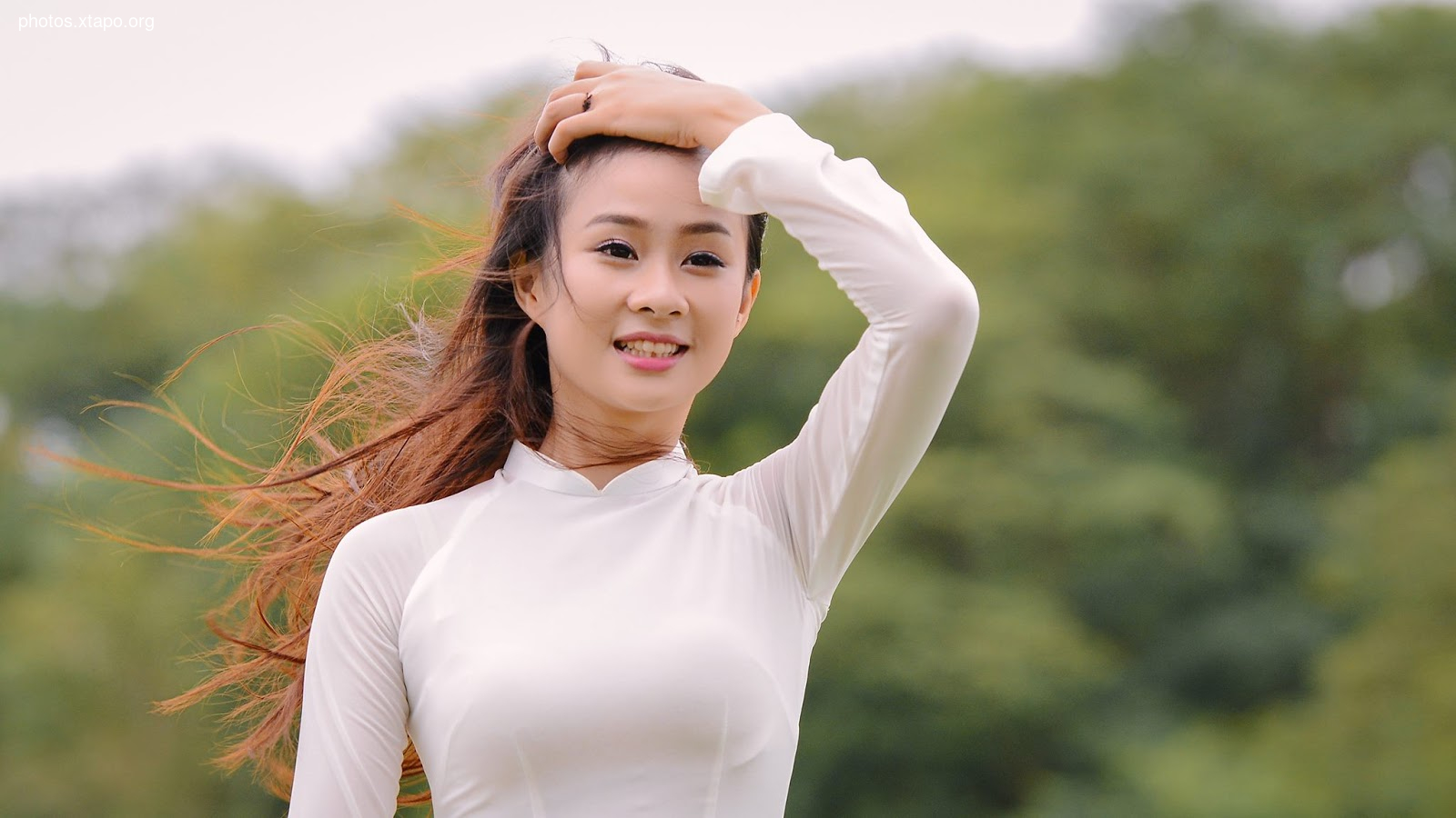 A young woman with long brown hair smiles gently as her hair blows in the breeze. She is wearing a traditional Vietnamese Ao Dai.