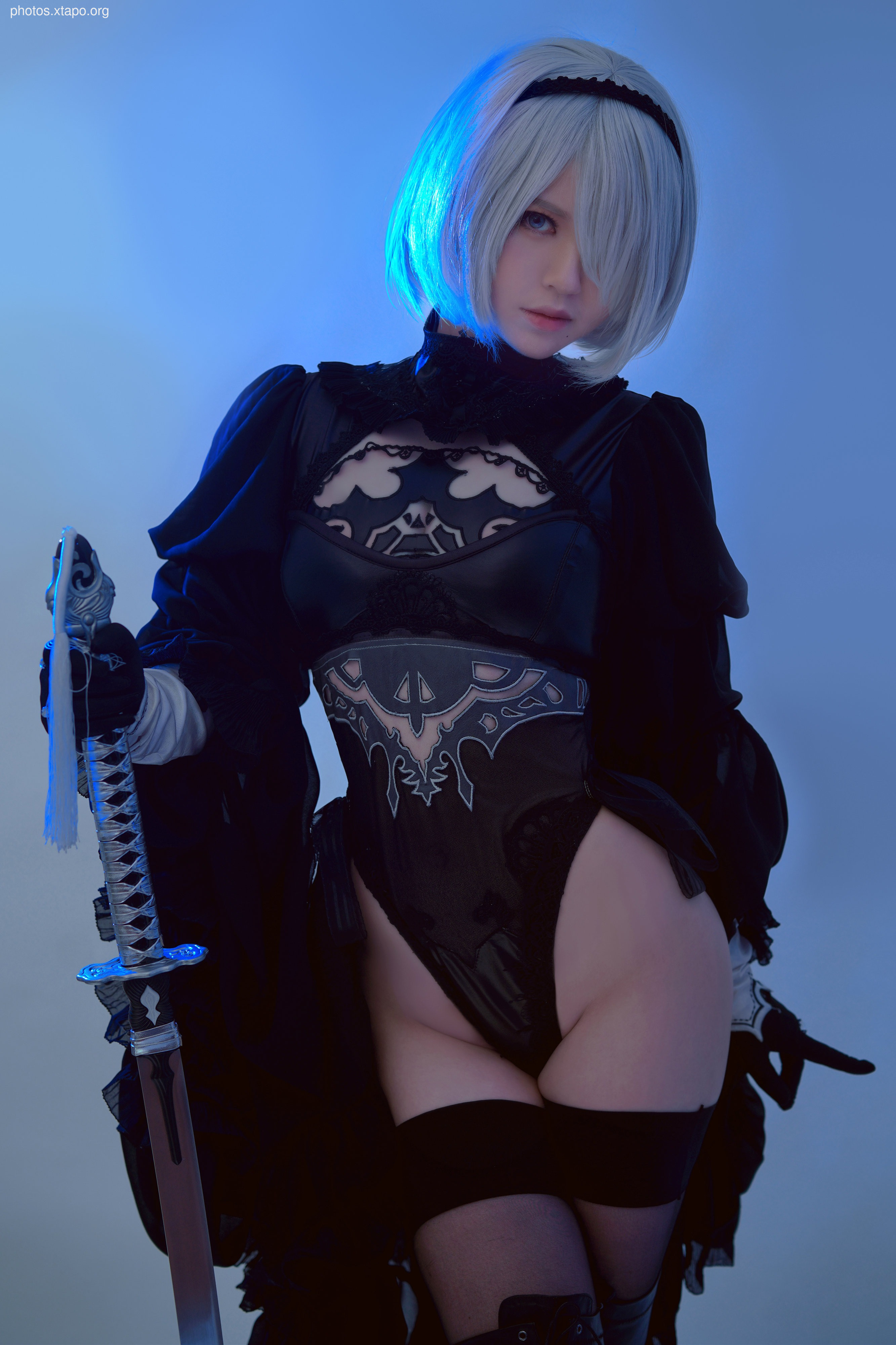 A stunning cosplay of 2B from Nier: Automata, wielding a katana and striking a powerful pose. The blue lighting enhances the overall aesthetic.