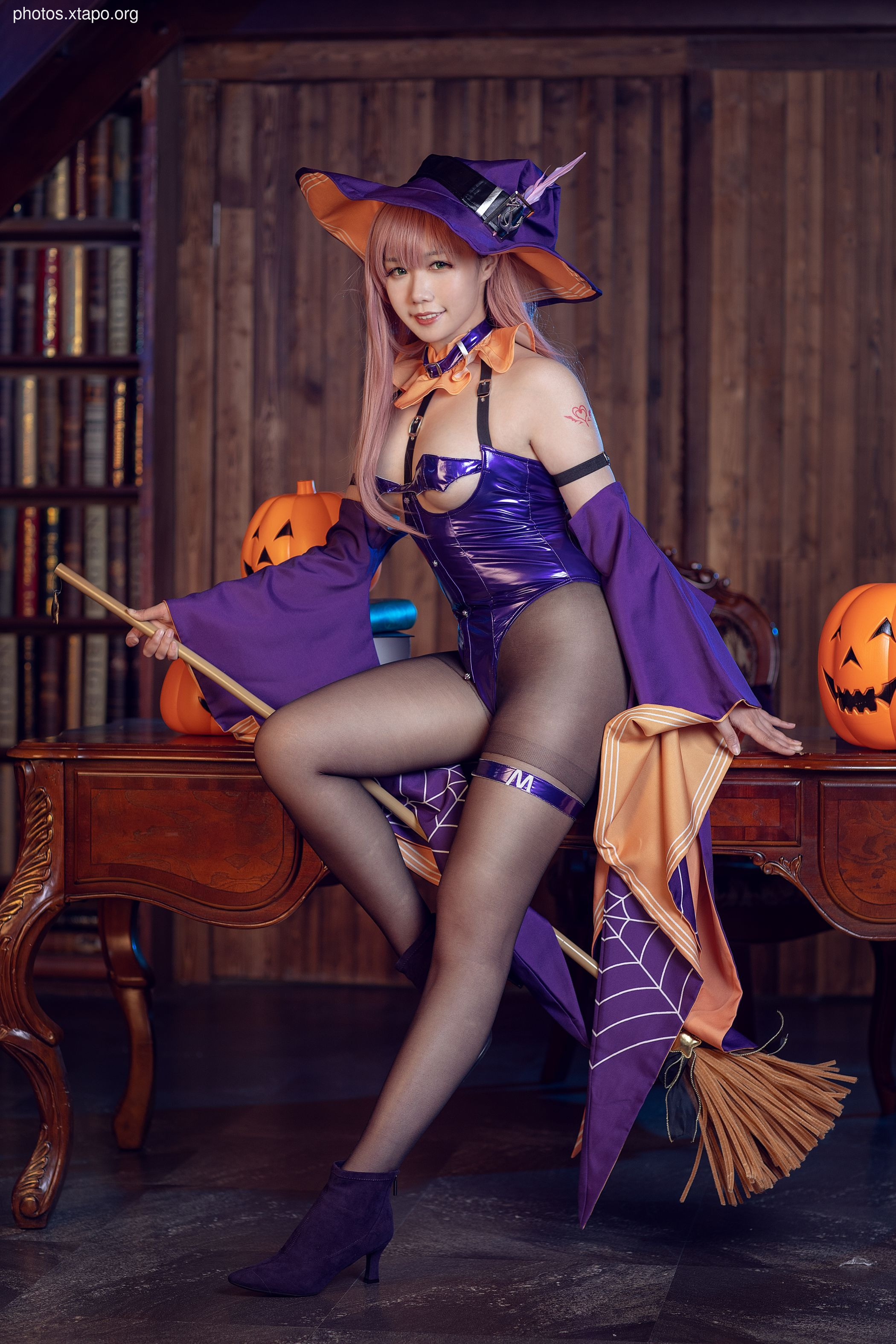 A captivating cosplay model, dressed as a Halloween witch, poses with a broom.  She's wearing a purple and orange costume with a witch's hat and sits on a wooden table near pumpkins.