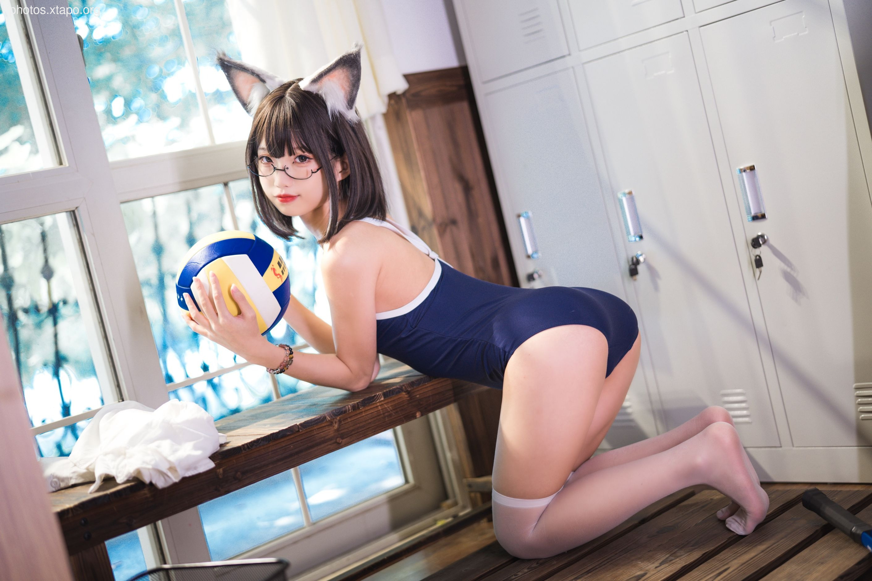 A young woman in a swimsuit holds a volleyball. She is wearing glasses and cat ears. The setting appears to be a locker room.