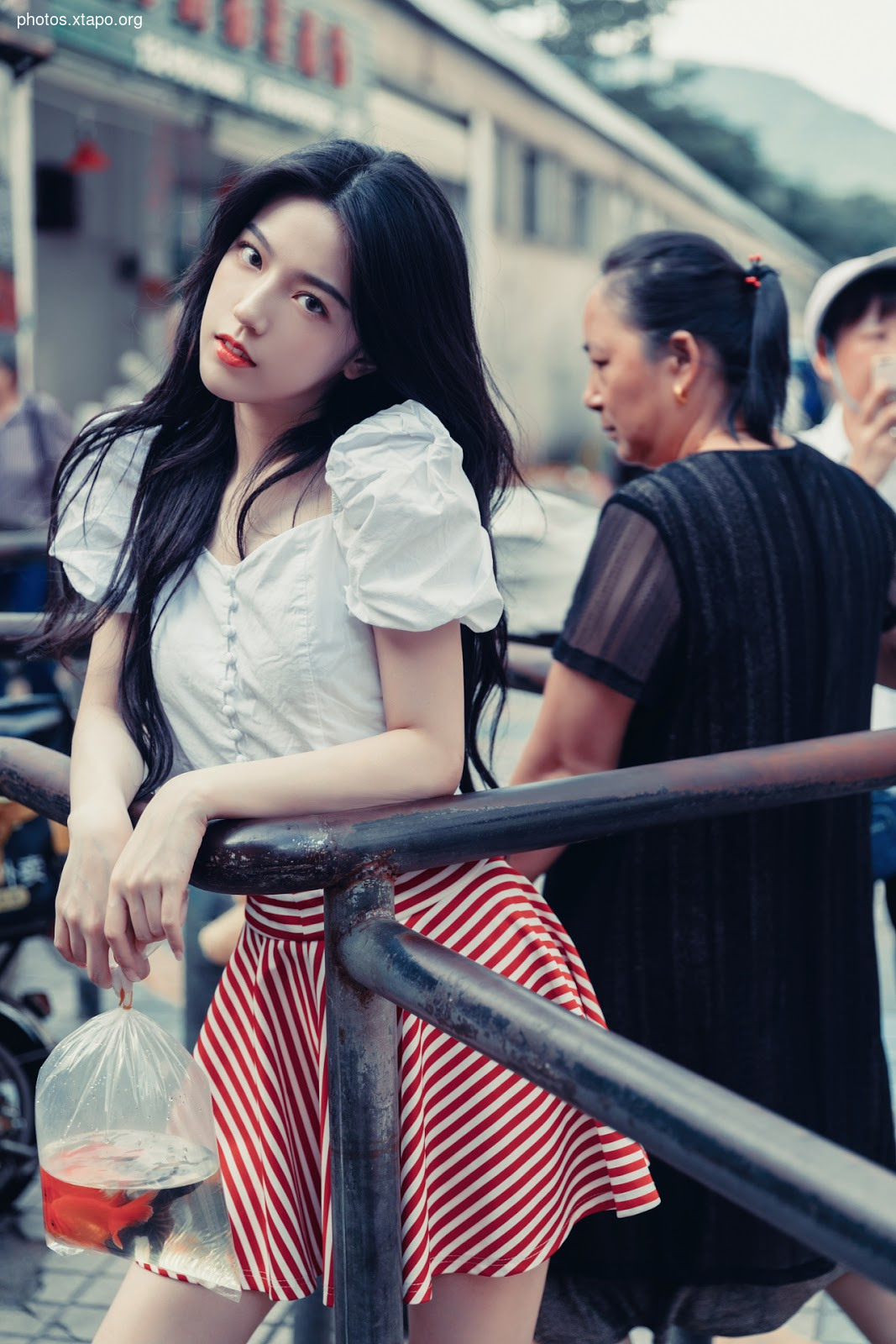 A young woman with long black hair stands on a street, holding a plastic bag containing a goldfish. She wears a white top and a red and white striped skirt. Behind her, another woman and a man are visible.