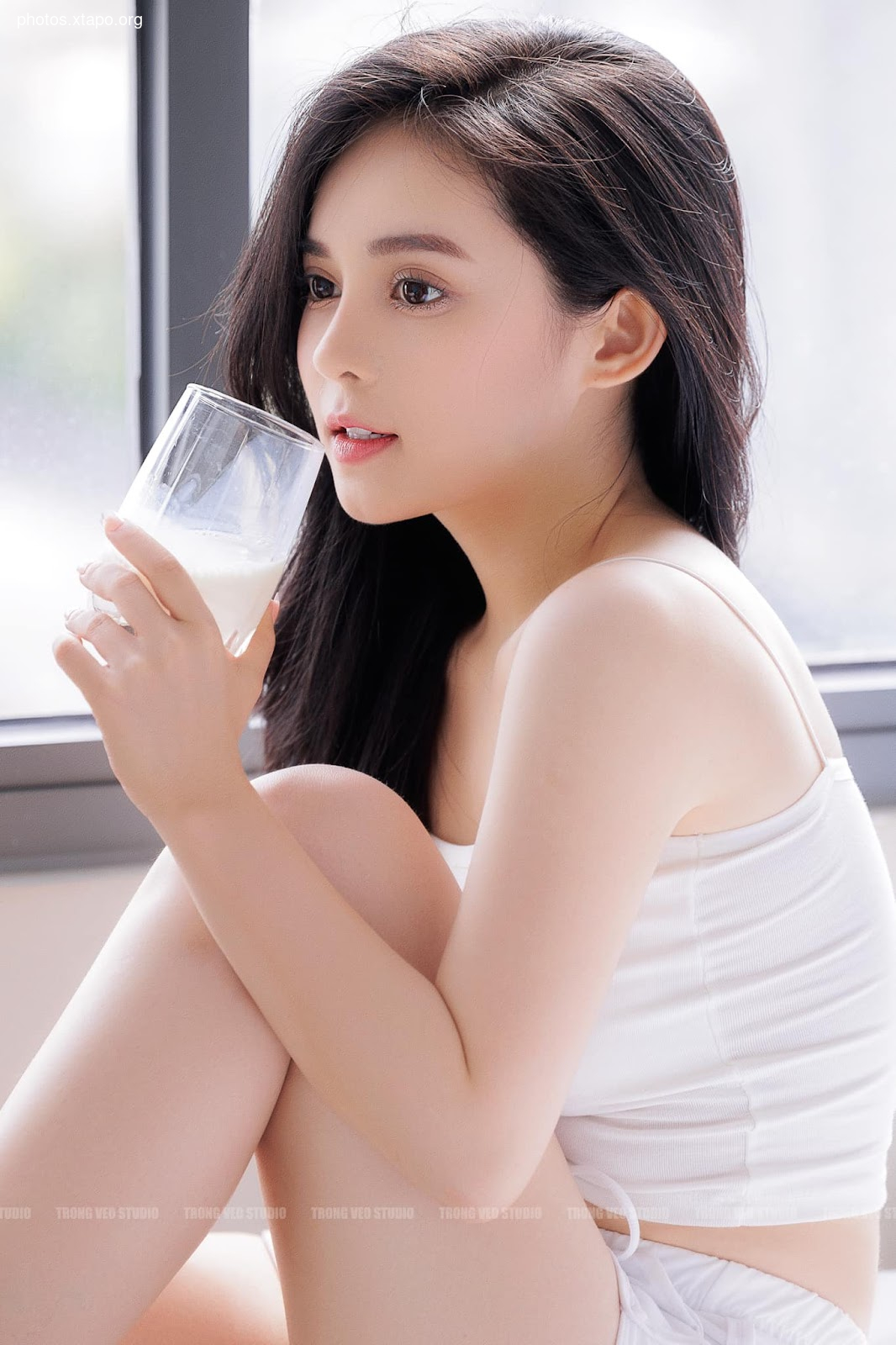 A young woman with long dark hair sits by a window, enjoying a refreshing glass of milk. She is wearing a white tank top and looks peaceful and content.