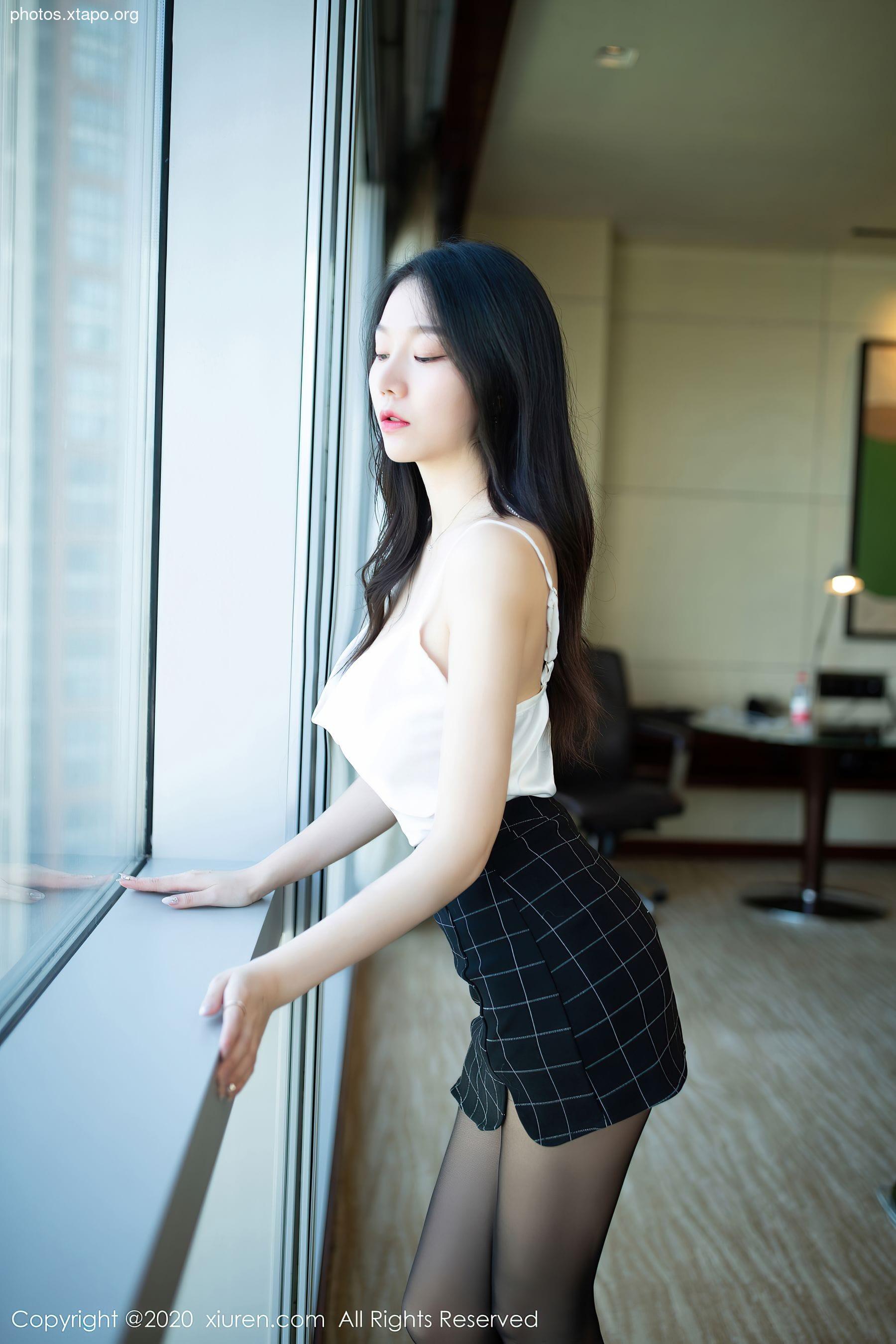 A captivating young woman gazes out a large window, dressed in a chic white top and a stylish black and white checkered skirt.