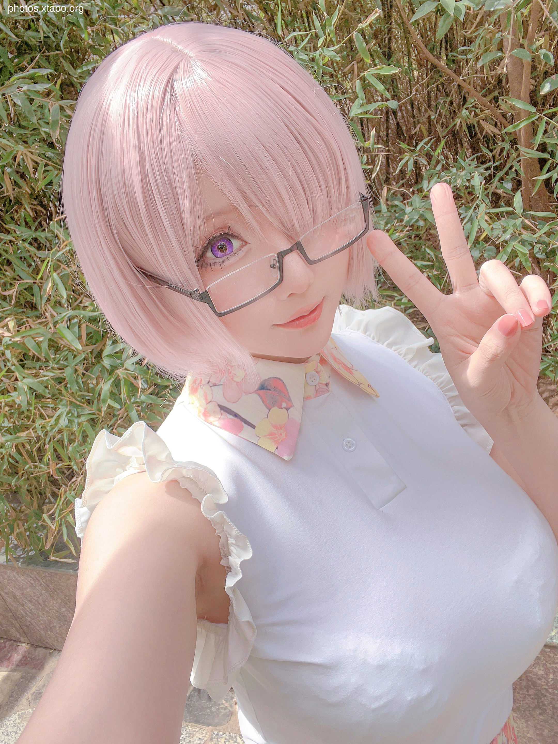 A cosplay of a girl with pink hair, wearing glasses and a white shirt with a flower-patterned collar. She is making a peace sign with her hand.