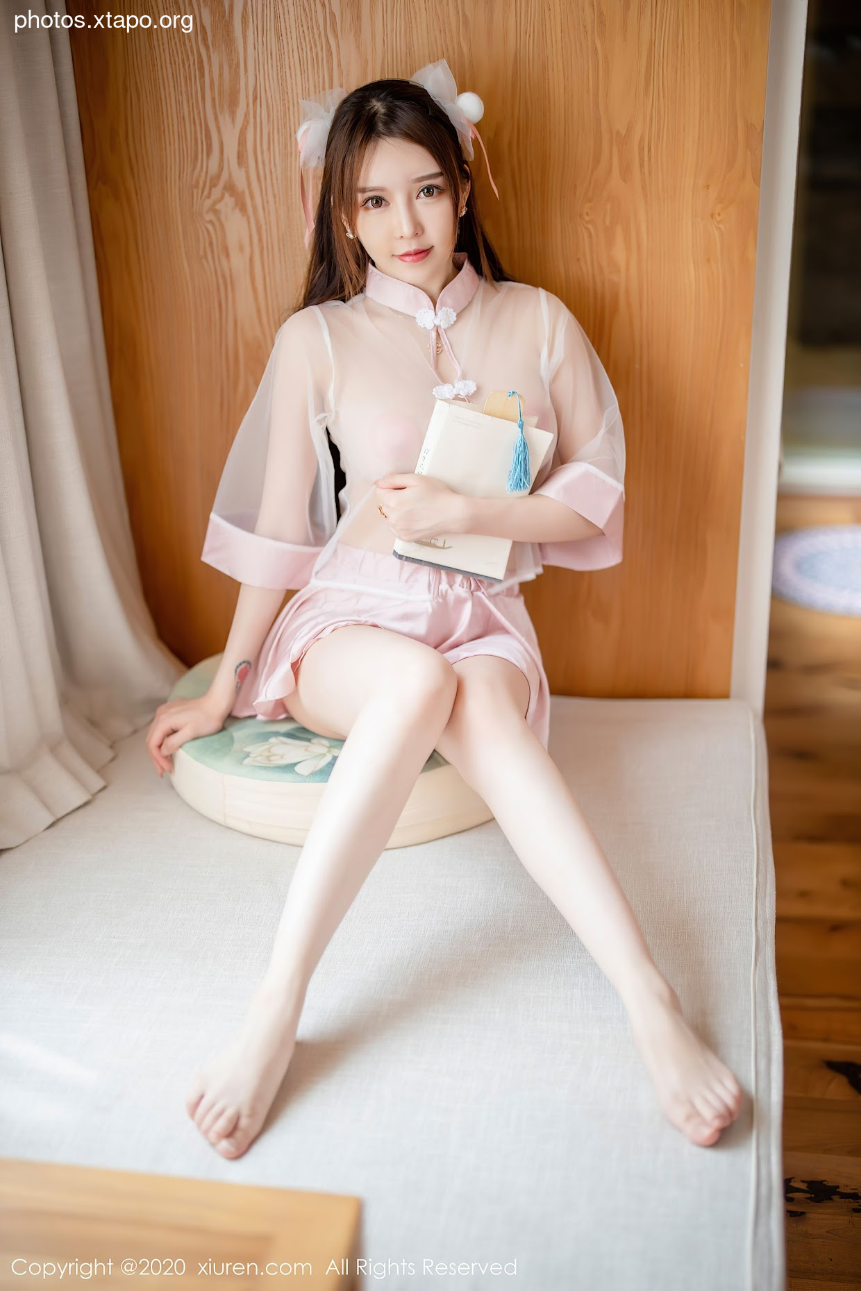 A young woman in a pink traditional Chinese dress sits on a floor cushion, holding a book.  She is wearing a cute hair accessory.