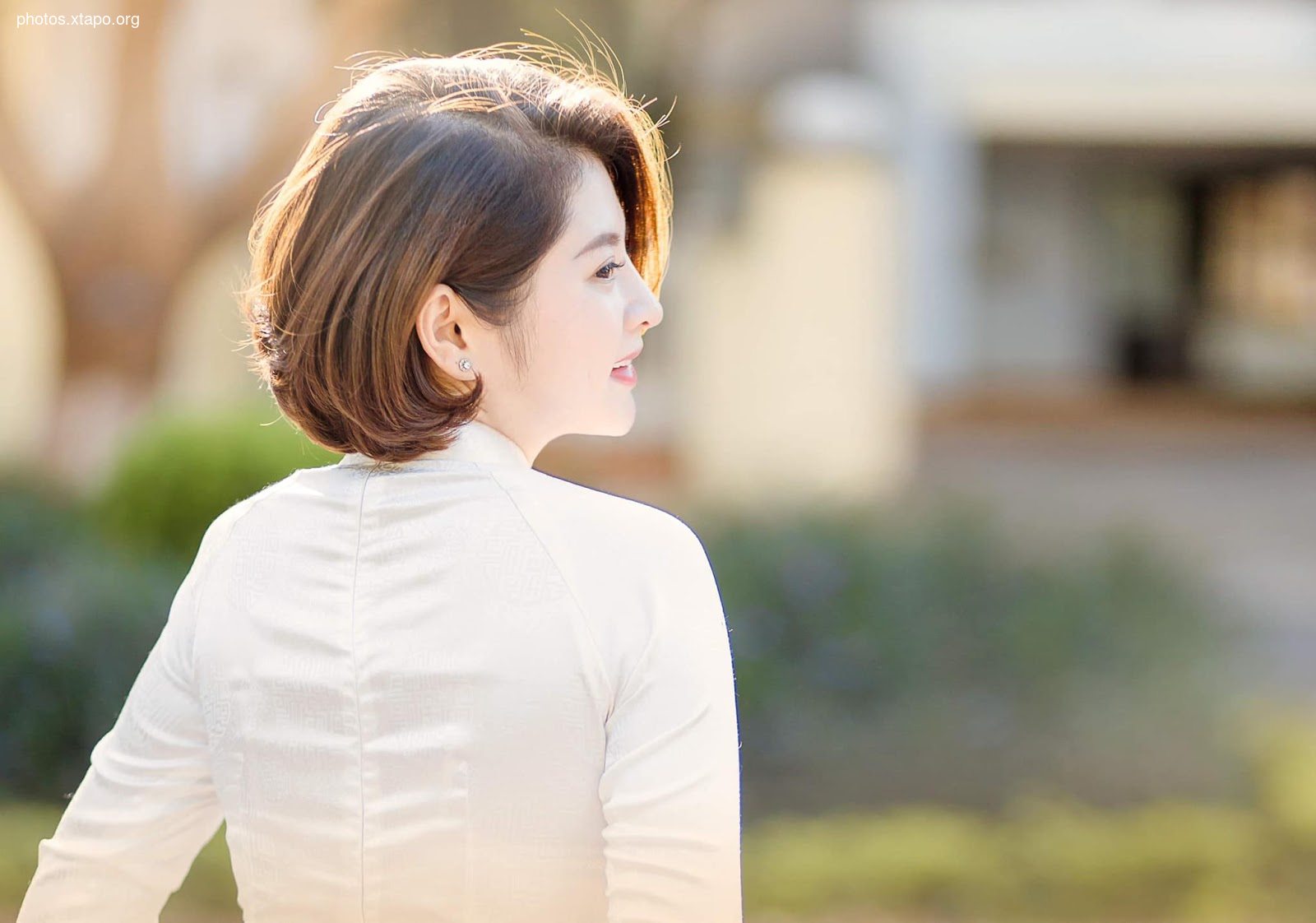 A young woman with short brown hair, wearing a traditional Vietnamese white Ao Dai, turns her head slightly away from the camera. The background is softly blurred, suggesting a peaceful outdoor setting.