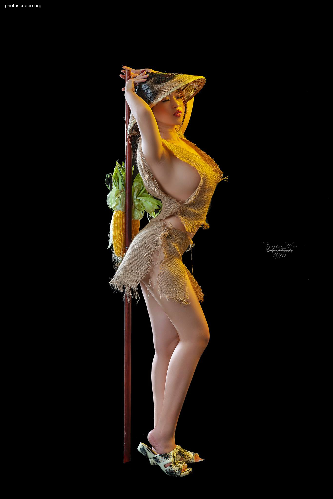 A woman wearing a burlap dress and conical hat stands holding a long pole with corn.  The background is black.