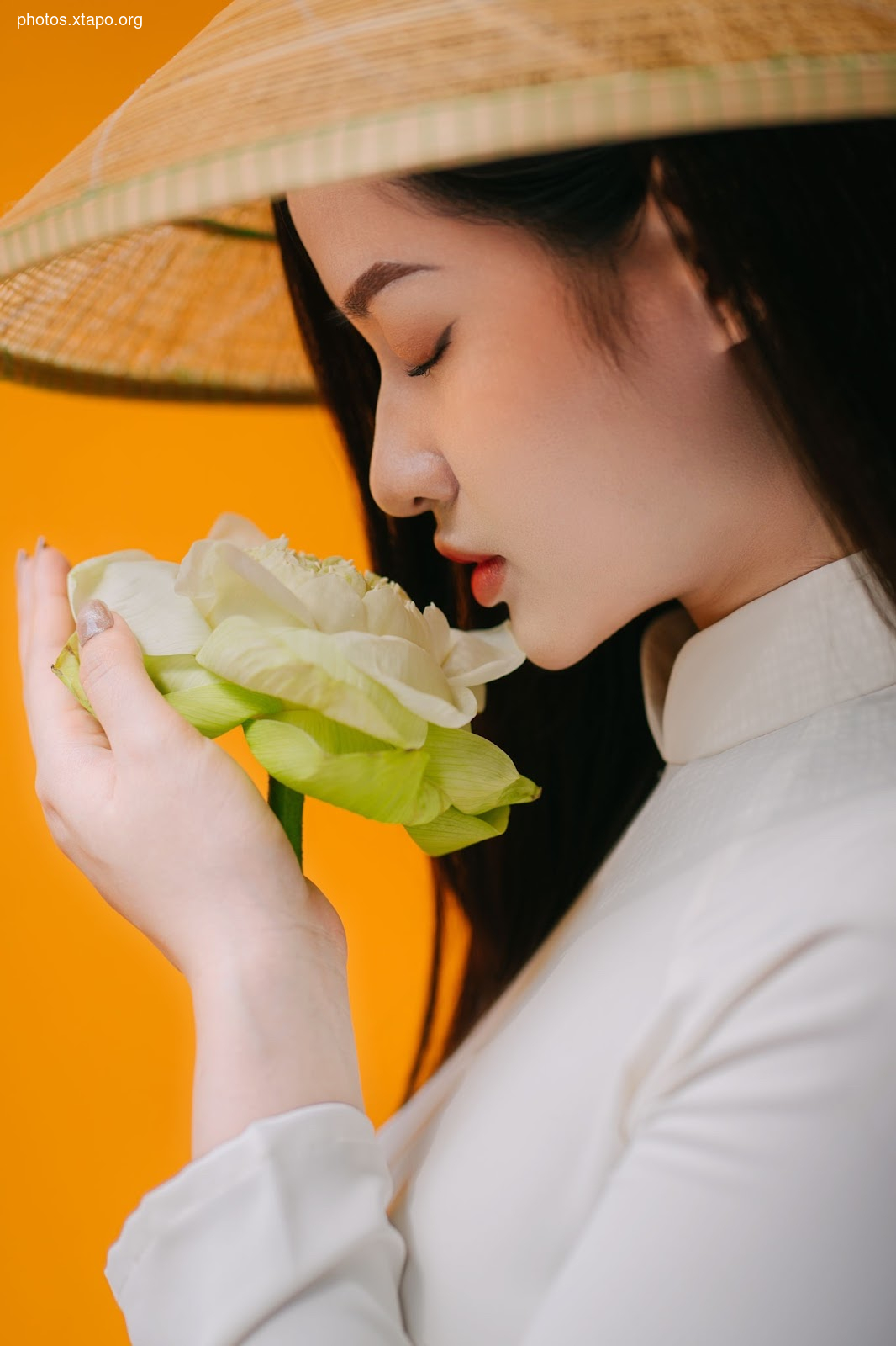 A young woman in traditional Vietnamese clothing gently smells a lotus flower. The image is set against a vibrant orange background.