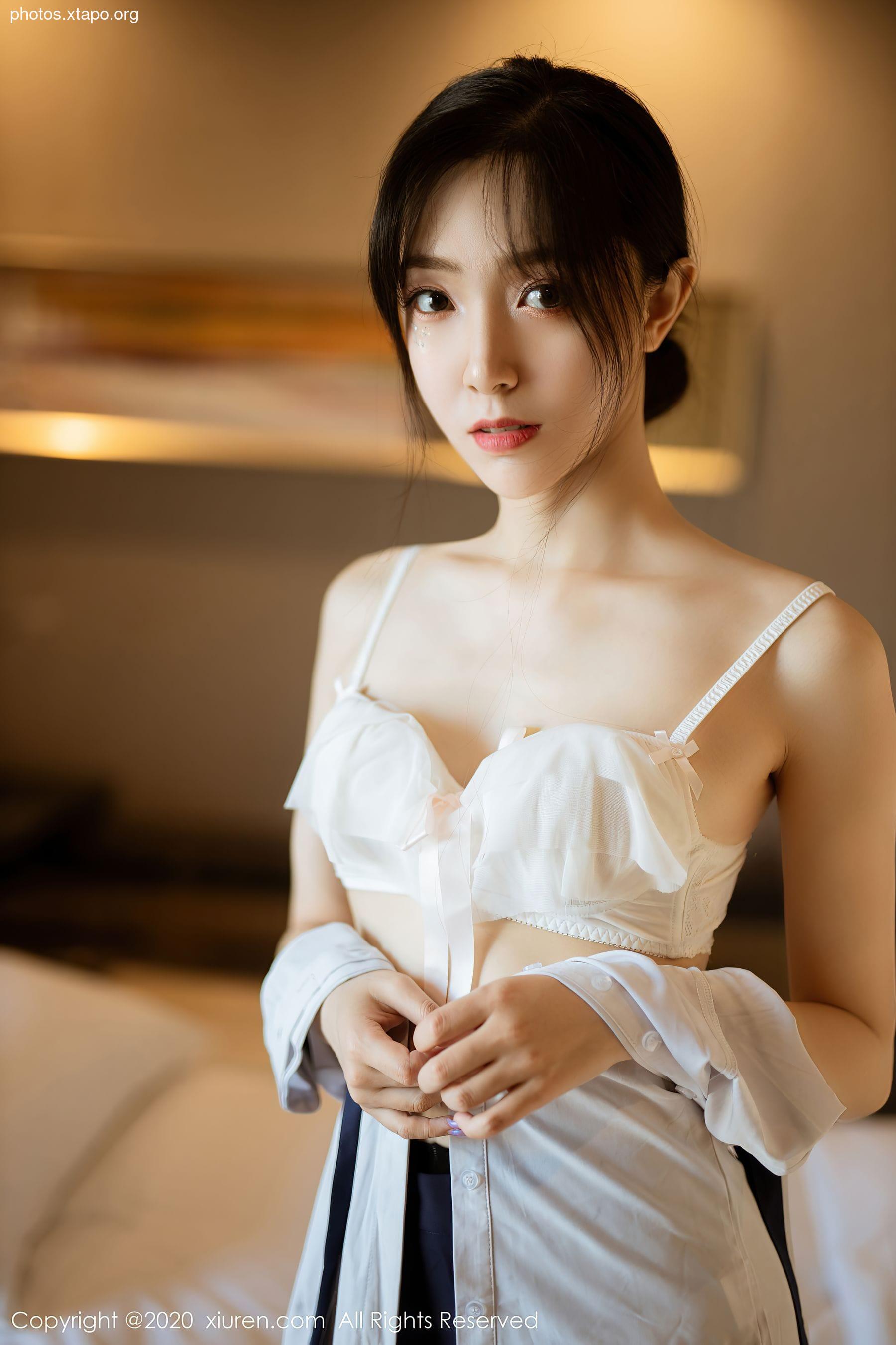 A young woman playfully adjusts her white shirt, revealing a hint of her lingerie beneath. The soft lighting and intimate setting create a sensual mood.