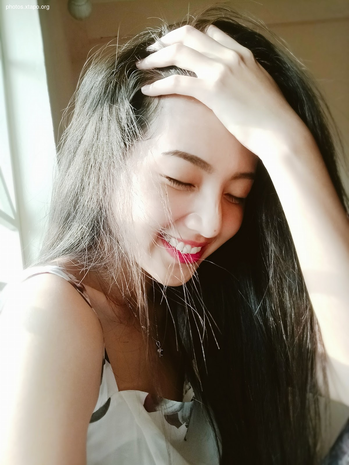 A young woman smiles gently, touching her hair.  She's wearing a white top.