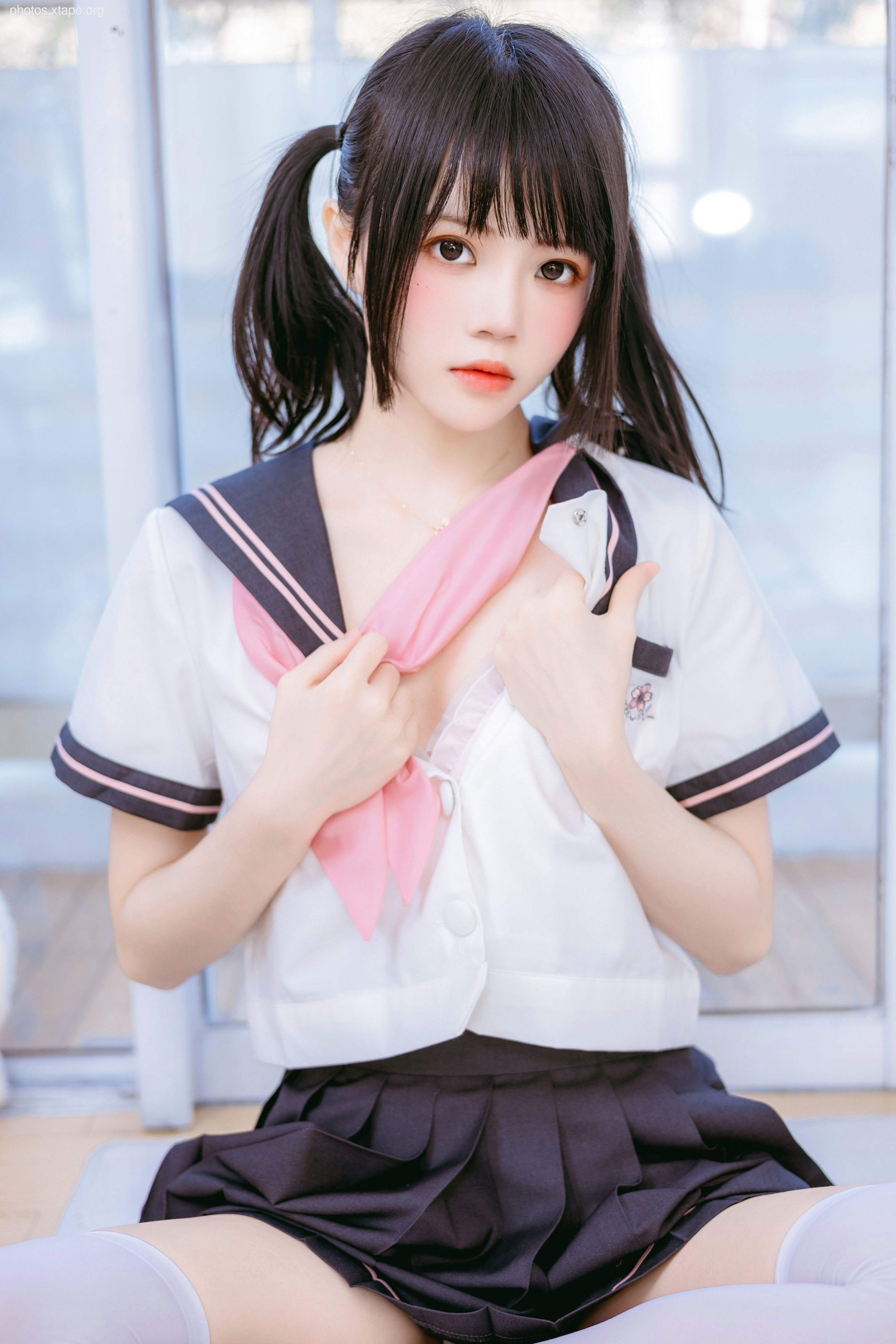 A young woman in a sailor school uniform poses for a photograph. She has her hair in pigtails and is wearing a pink scarf. The image is soft and dreamy.