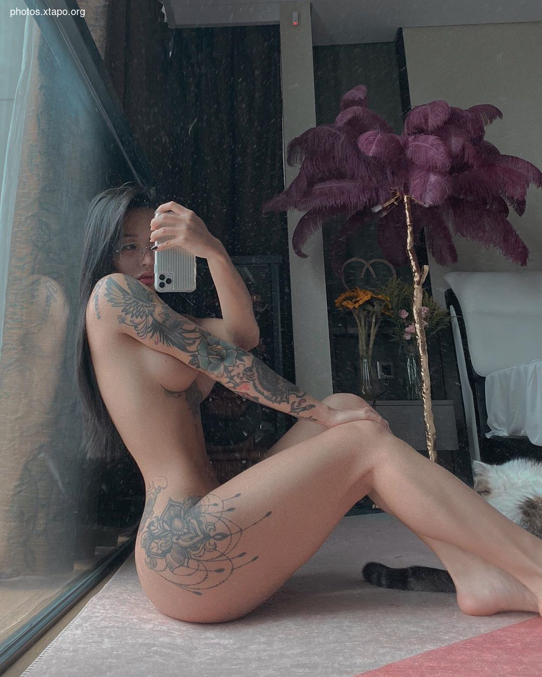 A young woman with extensive tattoos on her arms and legs takes a selfie in her apartment. She is sitting on the floor and a cat is visible in the background.