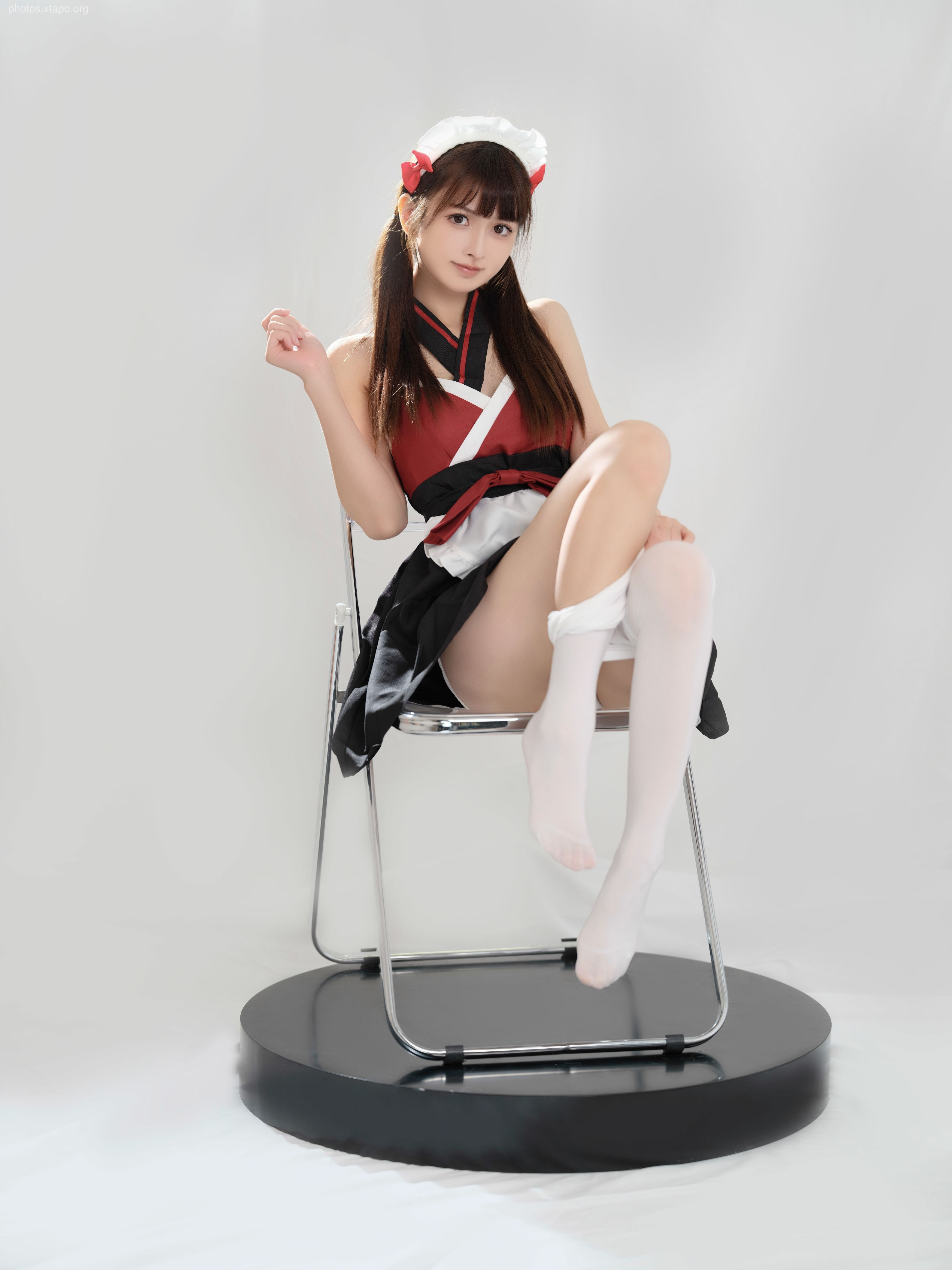 A young woman in a red and black cosplay outfit sits on a chair. She is wearing white socks and has her hair styled in pigtails.