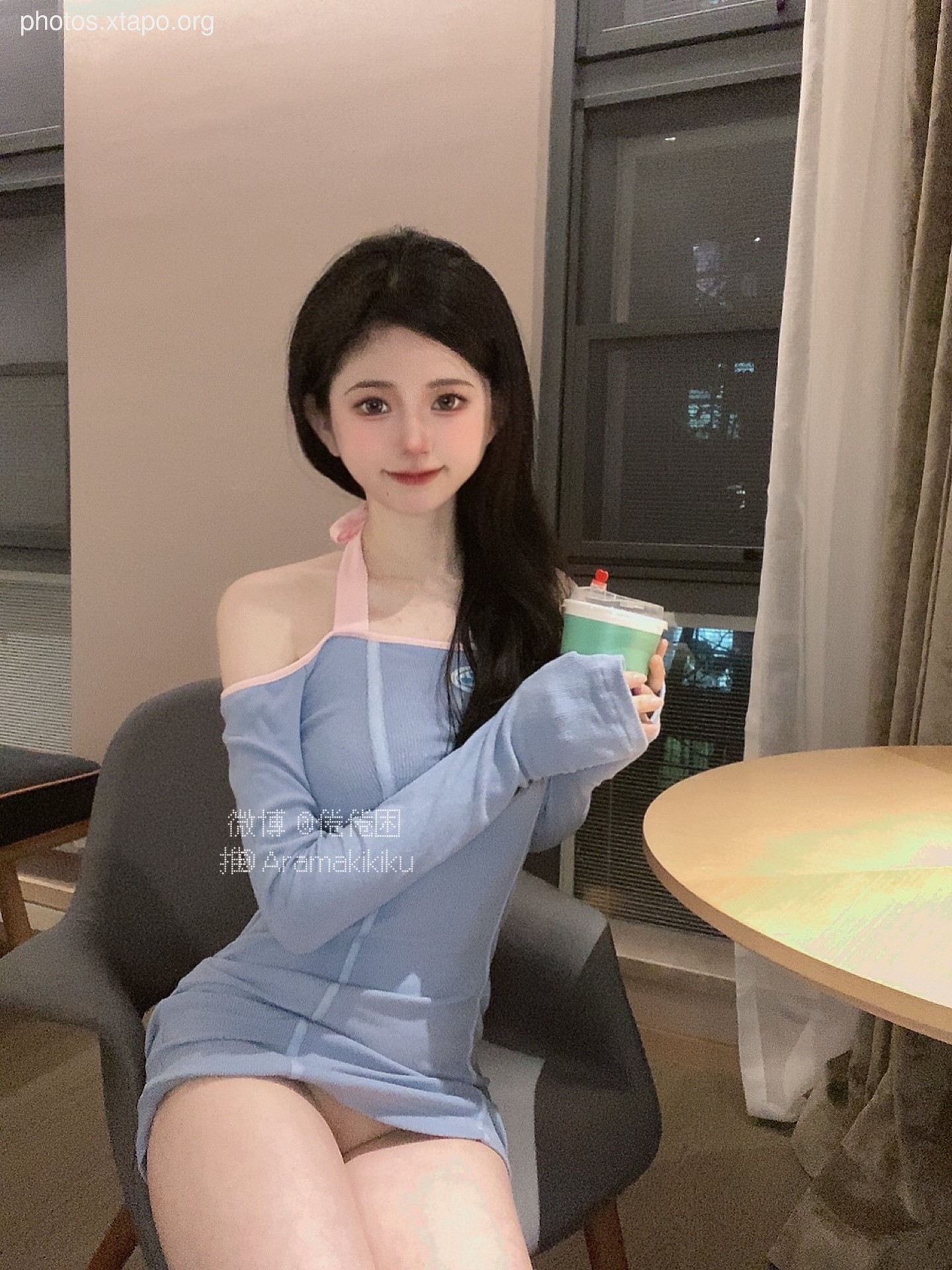 A young woman in a light blue off-the-shoulder dress sits at a table, holding a teal-colored drink. She is looking at the camera.