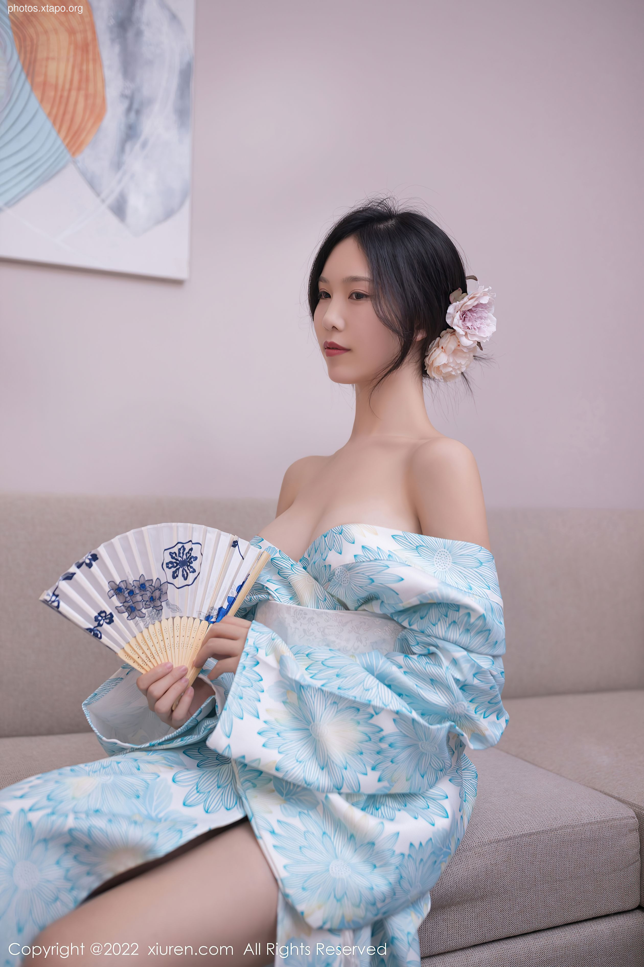 A young woman in a light blue yukata holds a hand fan.  She is sitting on a light gray couch.