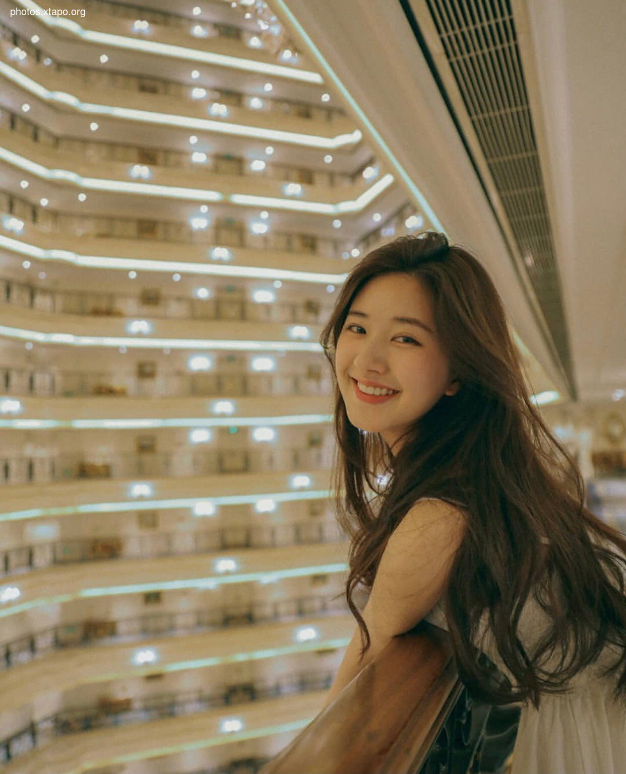 A young woman enjoys the evening view from a modern hotel. The building's interior is stunning, with warm lighting illuminating the many floors.