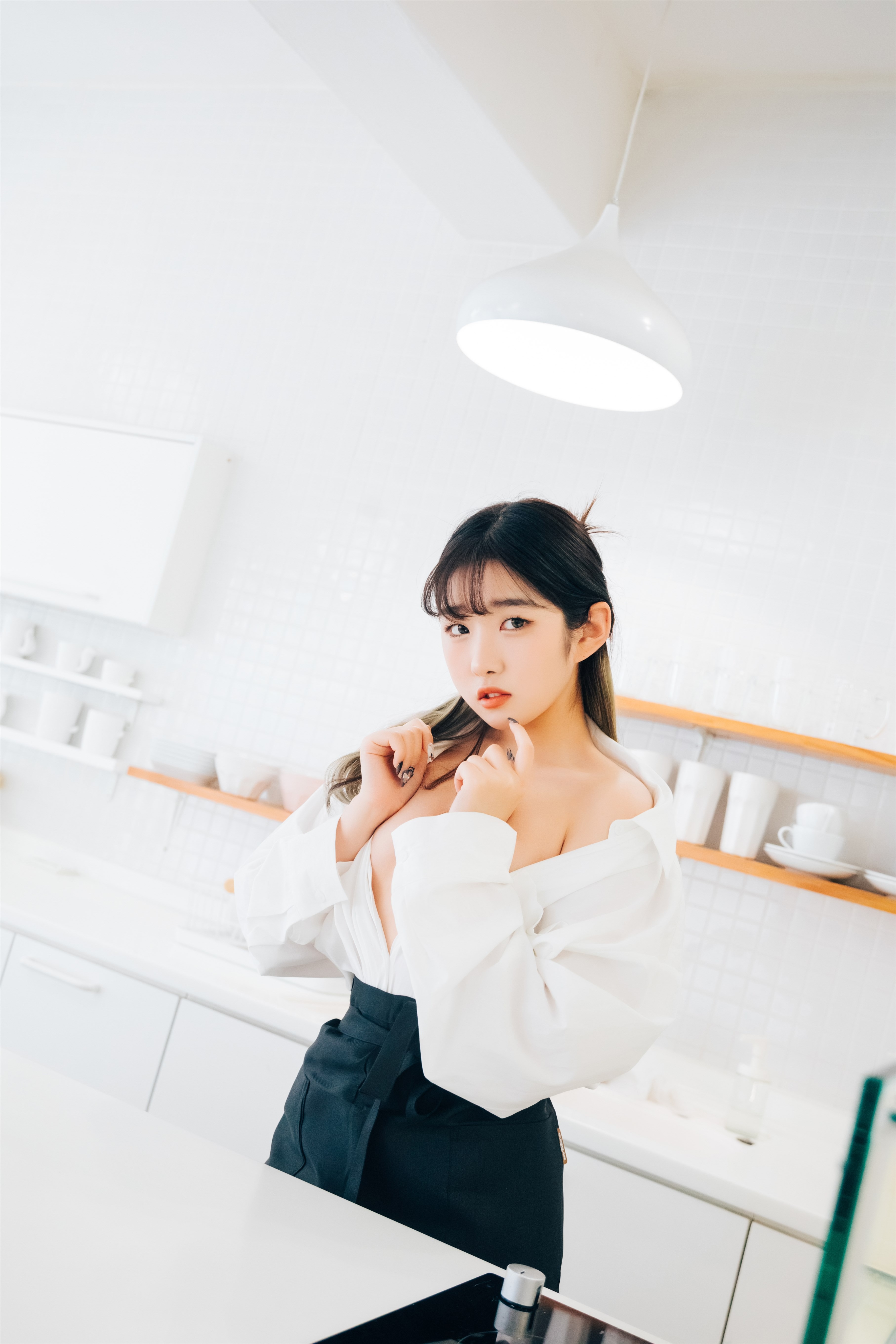 A young woman with dark hair stands in a bright white kitchen, wearing a white oversized shirt and a black skirt. She is slightly smiling and touching her shirt.