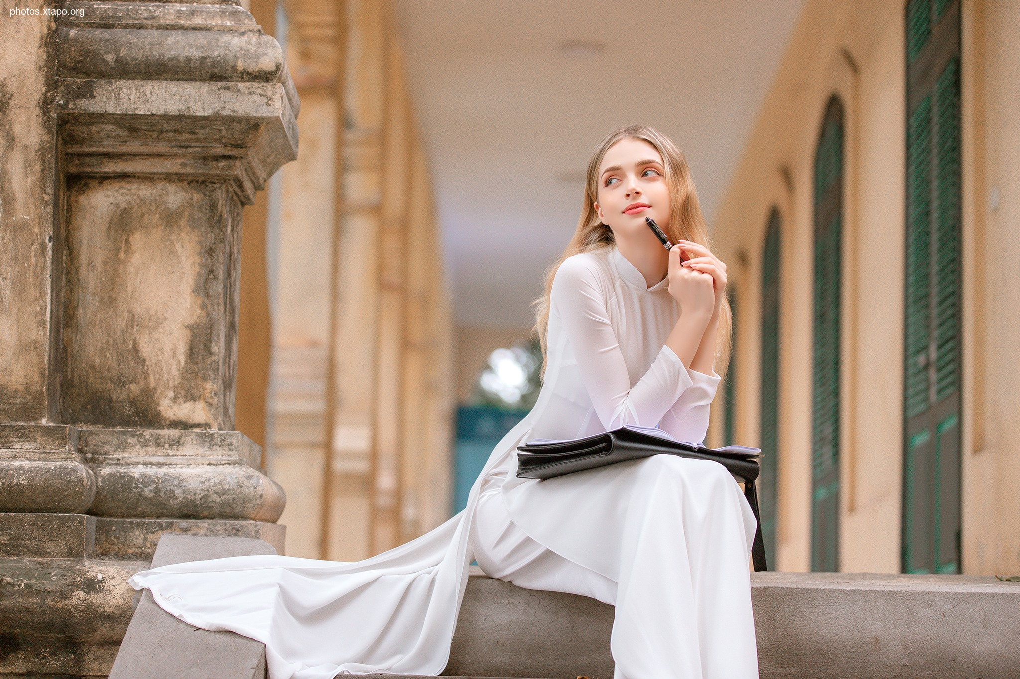 A young woman in a white ao dai sits on a stone step, pensively holding a pen and notebook, lost in thought.