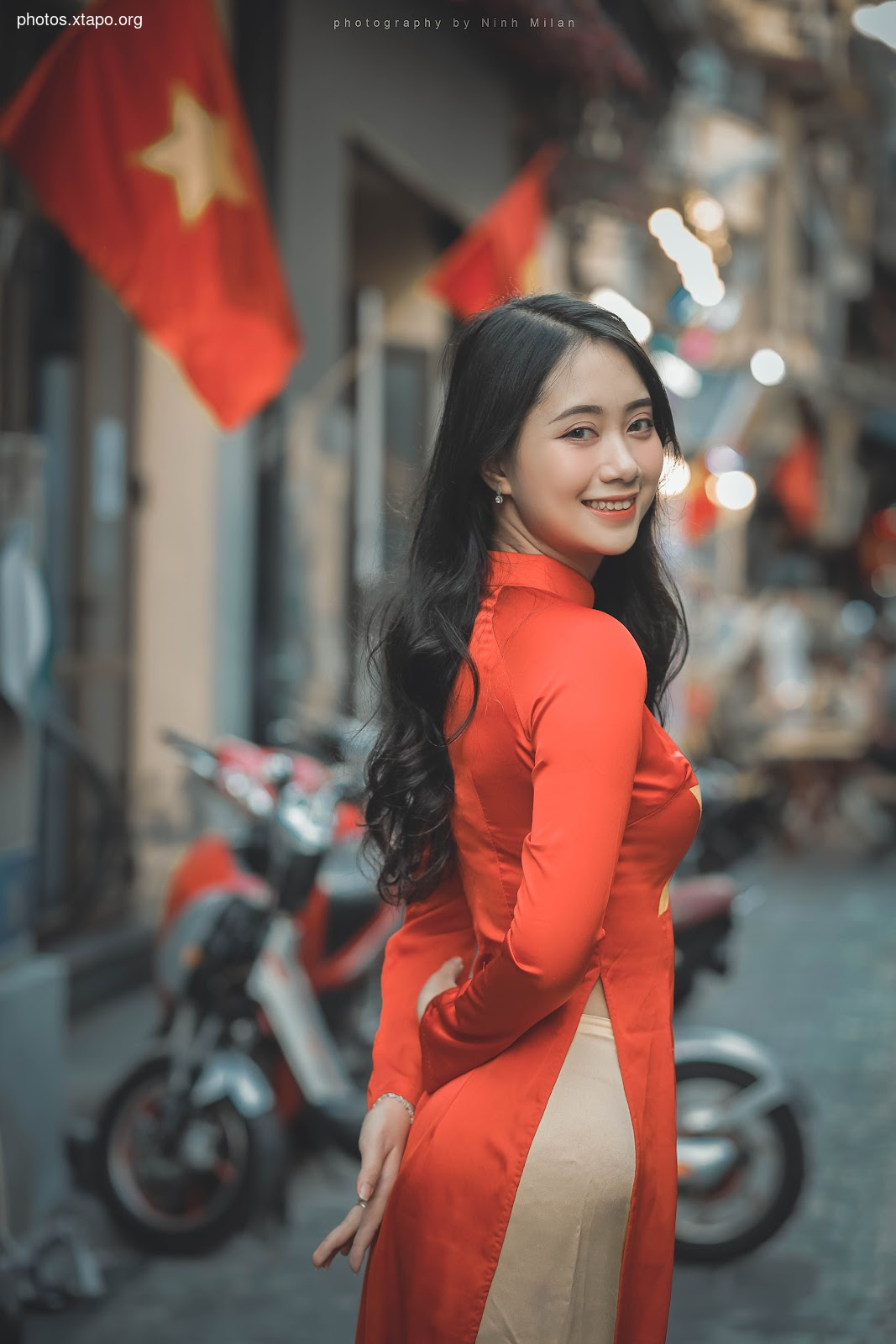 A young woman smiles while wearing a traditional Vietnamese Áo Dài dress in a bustling city street.