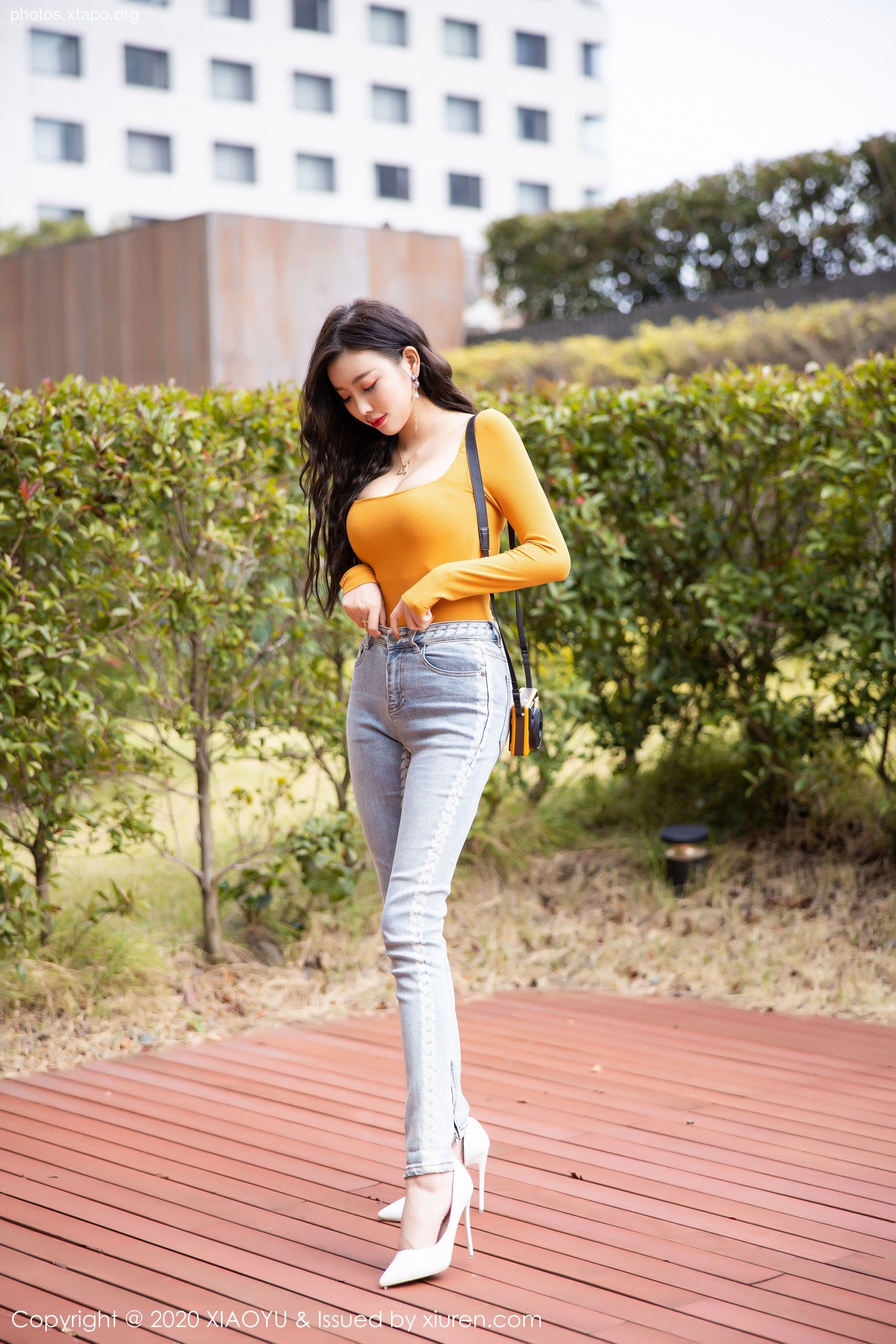 A fashionable young woman strolls outdoors, wearing a fitted yellow long-sleeve top, light-wash jeans, and white heels.  She carries a small handbag.
