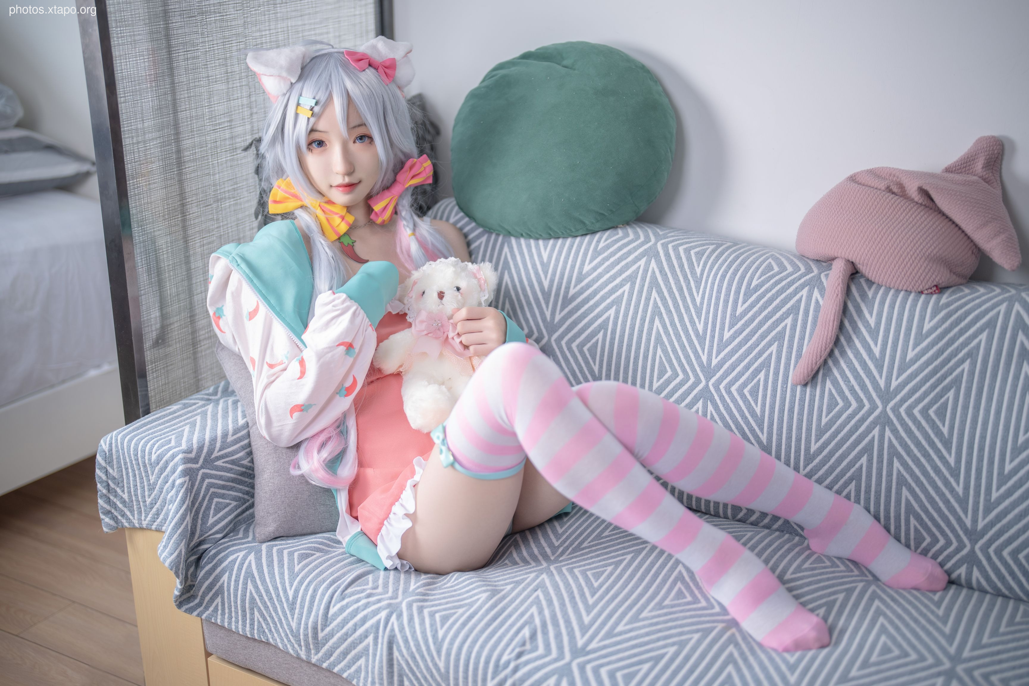 A young woman cosplaying in a pastel pink and mint green outfit with cat ears and a teddy bear. She's wearing striped knee-high socks and is sitting on a gray patterned couch.