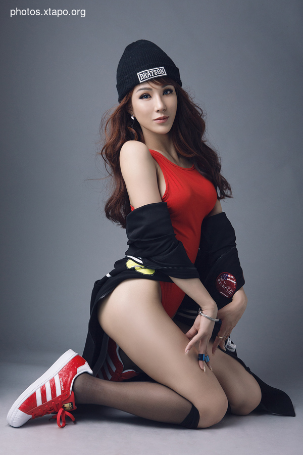 A fashion model poses in a red bodysuit, black beanie, and black jacket. She is wearing red Adidas sneakers and black stockings. The background is gray.