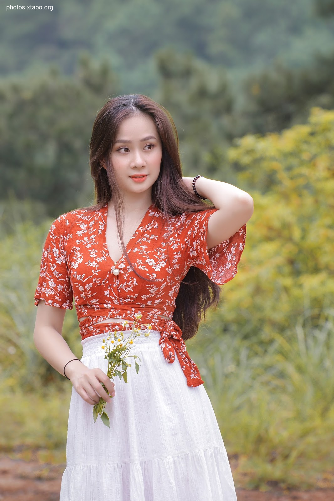 A young woman poses for a photograph in a red floral crop top and white skirt. She is holding a bouquet of flowers and smiling gently. The background is lush greenery.