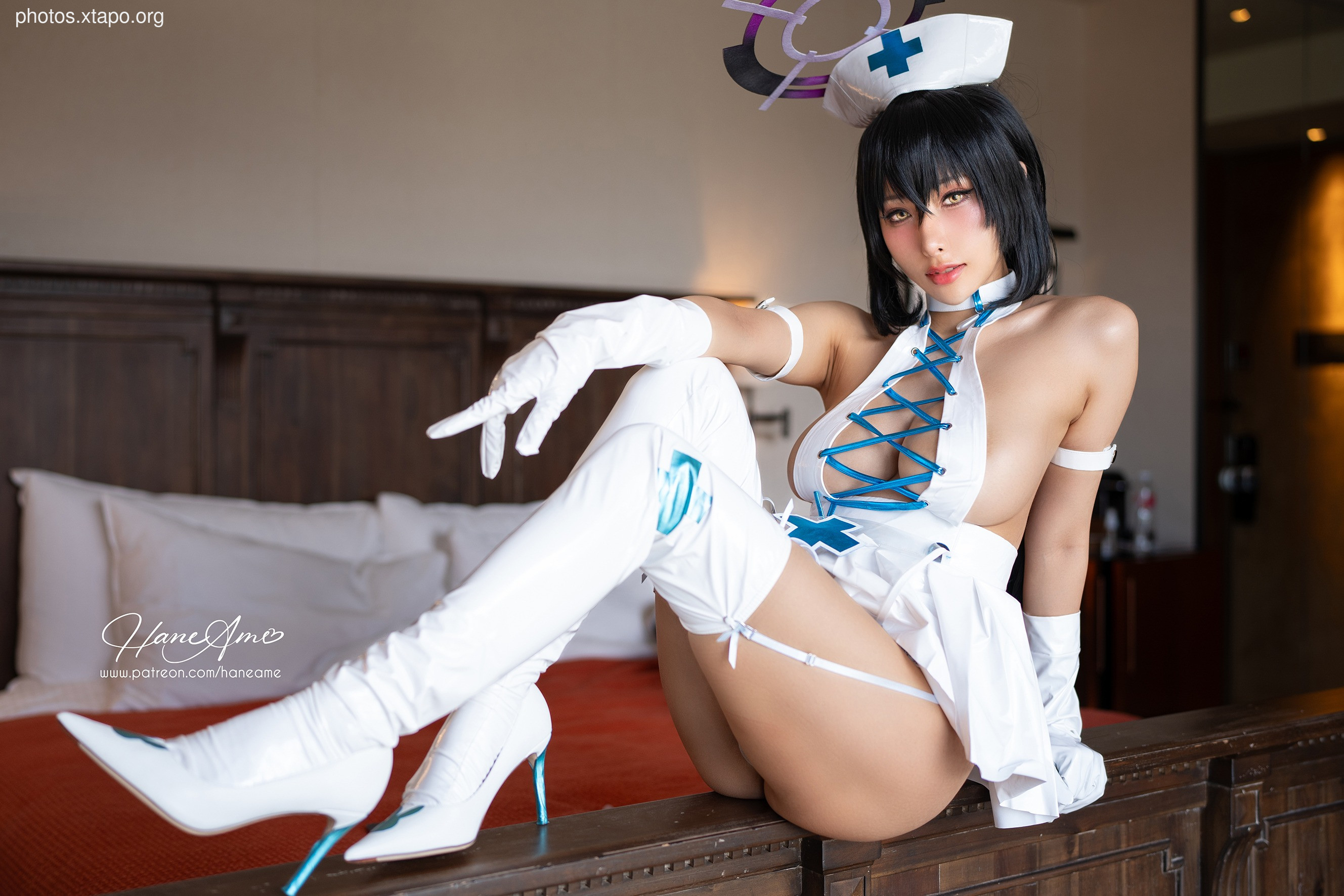 A model in a nurse cosplay sits on a bed in a hotel room.  She is wearing a white nurse's uniform with blue accents and white thigh-high boots.
