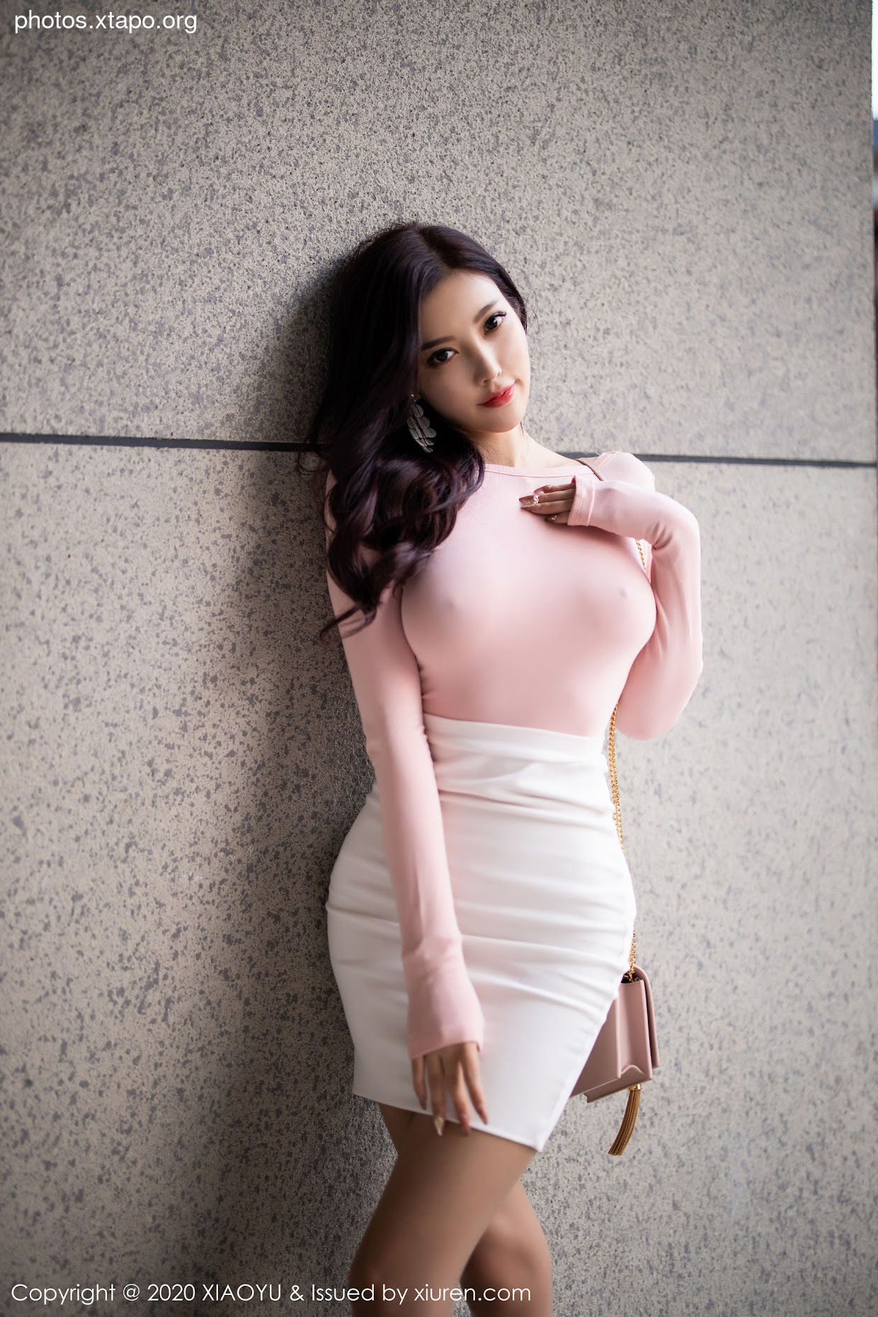 A woman in a pink long-sleeve top and white skirt poses against a textured wall.