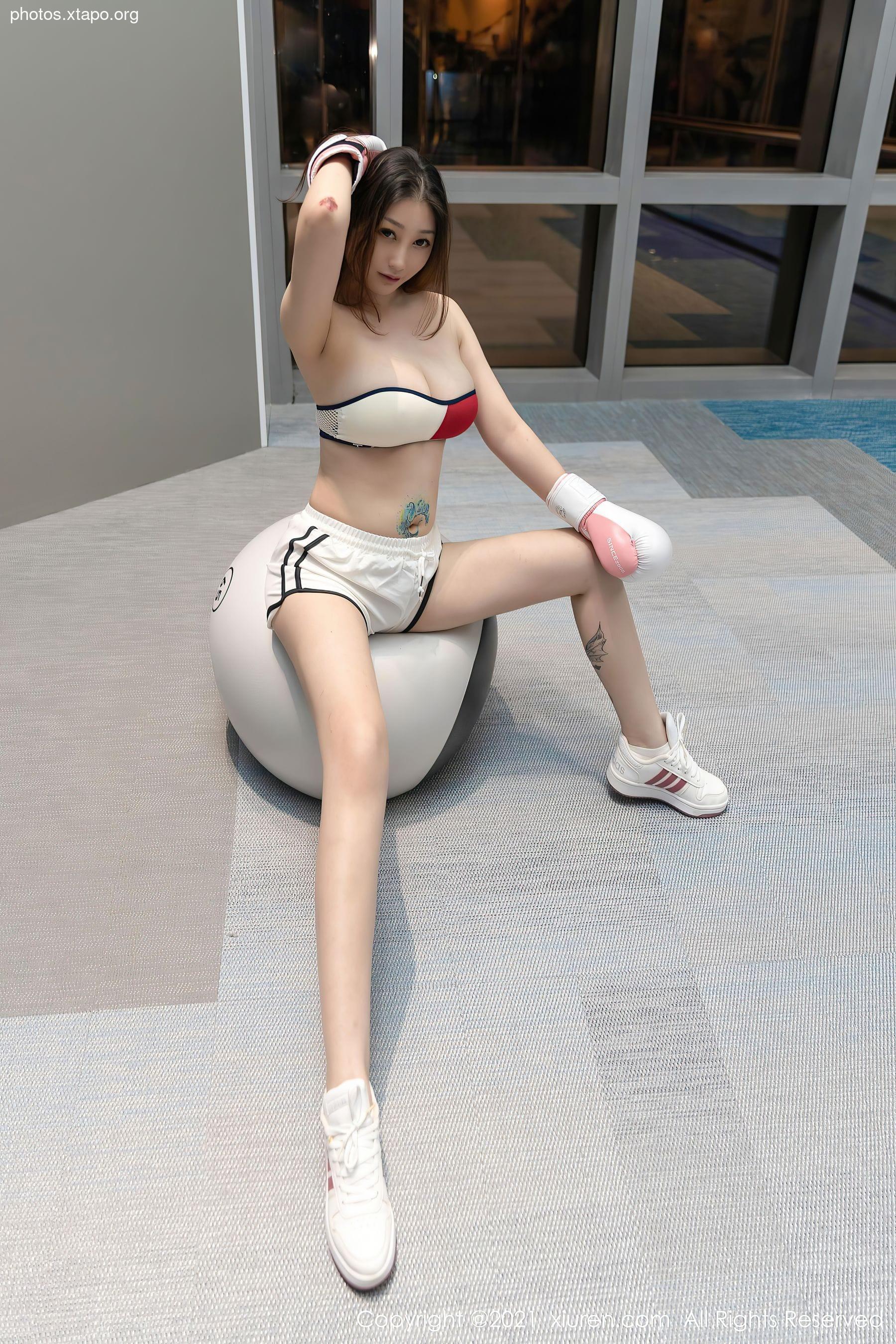 A young woman in a white sports bra and shorts sits on an exercise ball, wearing boxing gloves.