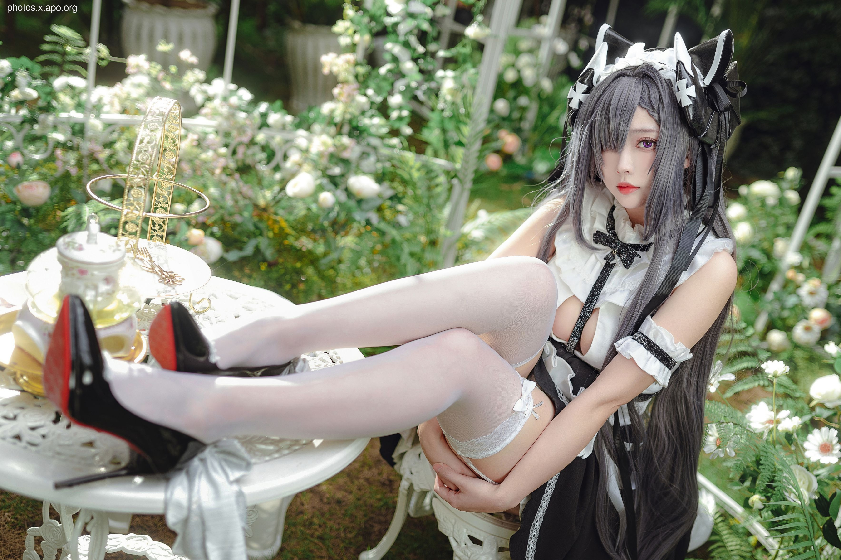 A young woman in a maid cosplay sits in a garden setting, wearing a black and white dress and cat ears. She is posing for a photo.