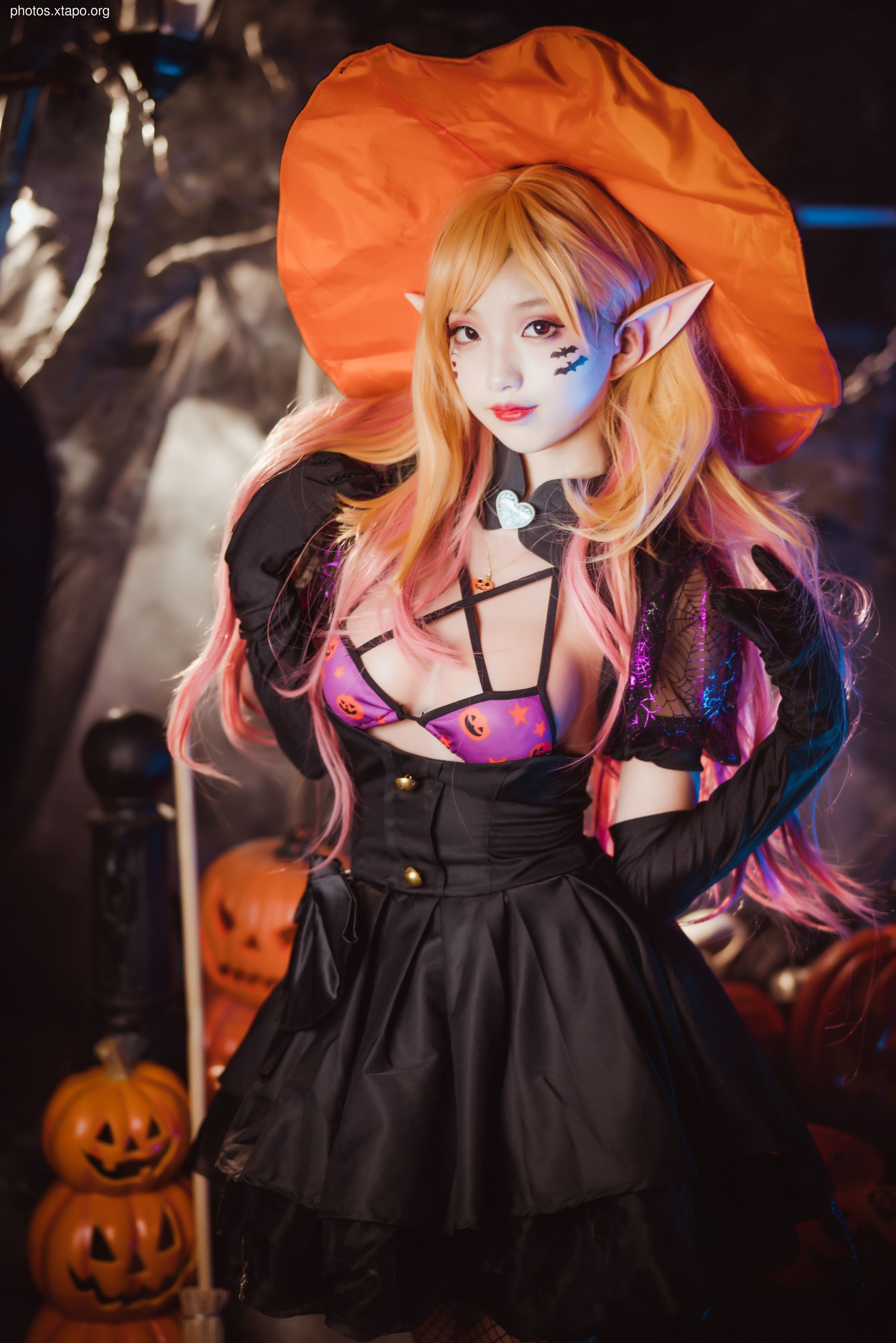 A young woman in a gothic lolita witch costume poses for a Halloween photoshoot. She is wearing a black dress, a purple bra, and a large orange witch's hat. Pumpkins and Halloween decorations are in the background.