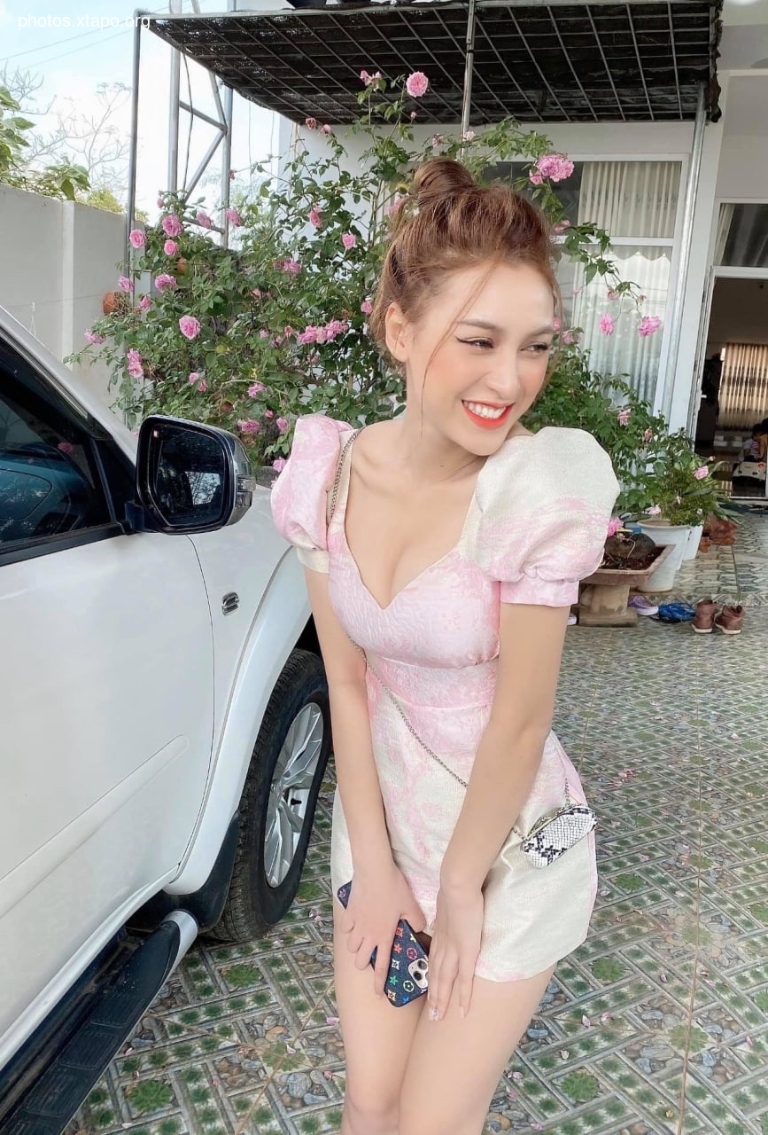 A young woman smiles while standing in front of a car wearing a pink dress.