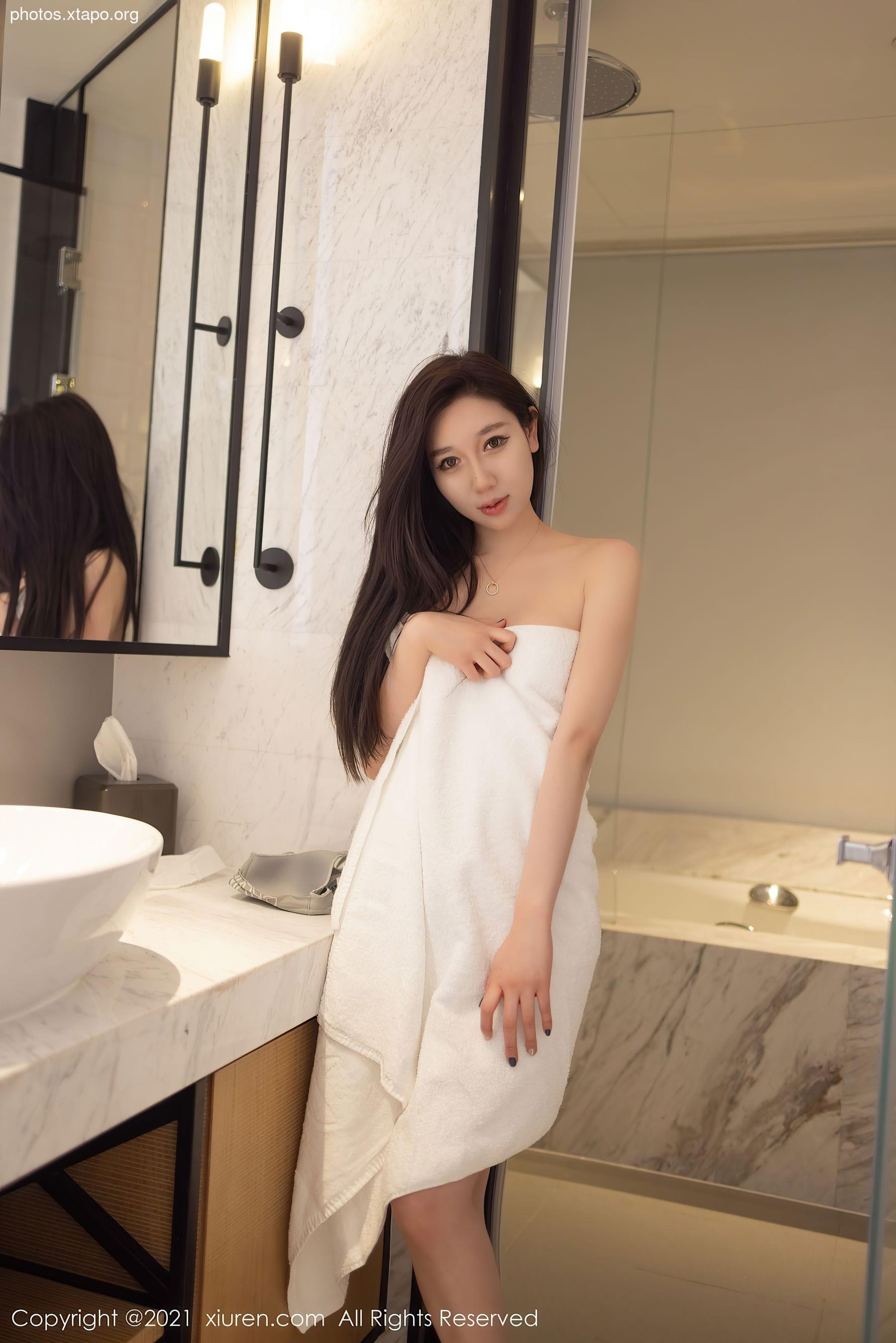 A young woman stands in a bathroom wrapped in a white towel.