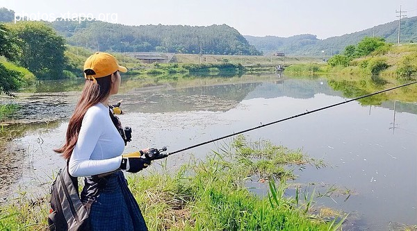 A woman wearing a yellow cap and white long-sleeve shirt enjoys a relaxing day of fishing at a calm lake surrounded by lush greenery and mountains.