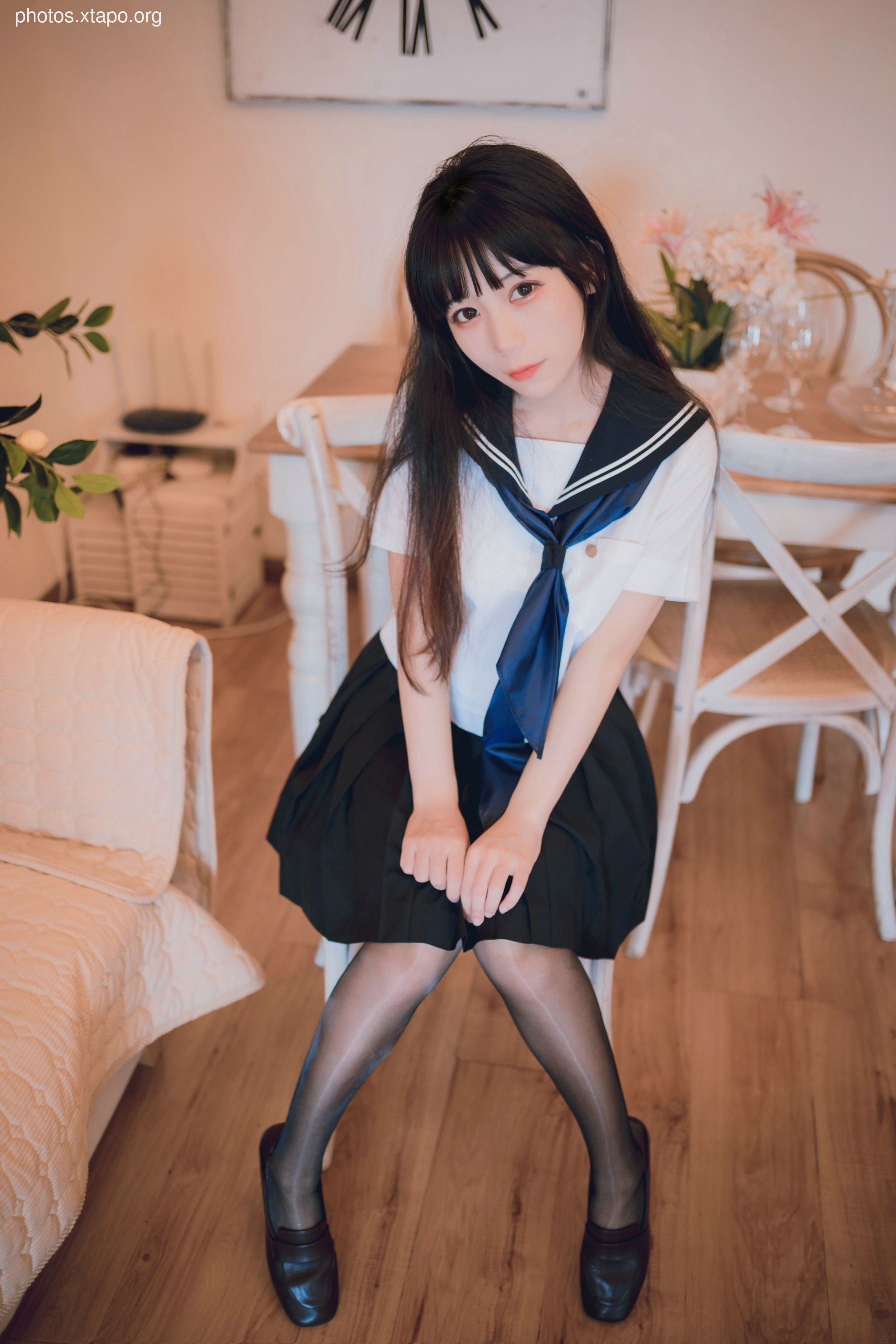 A young woman in a sailor school uniform sits on a chair, looking playfully mischievous.