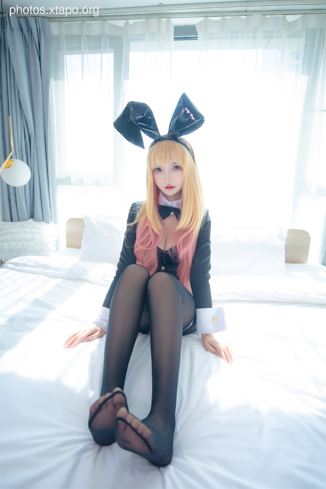 A young woman in a black bunny girl costume sits on a bed, posing for a photo.  She has long blonde hair and is wearing black stockings.