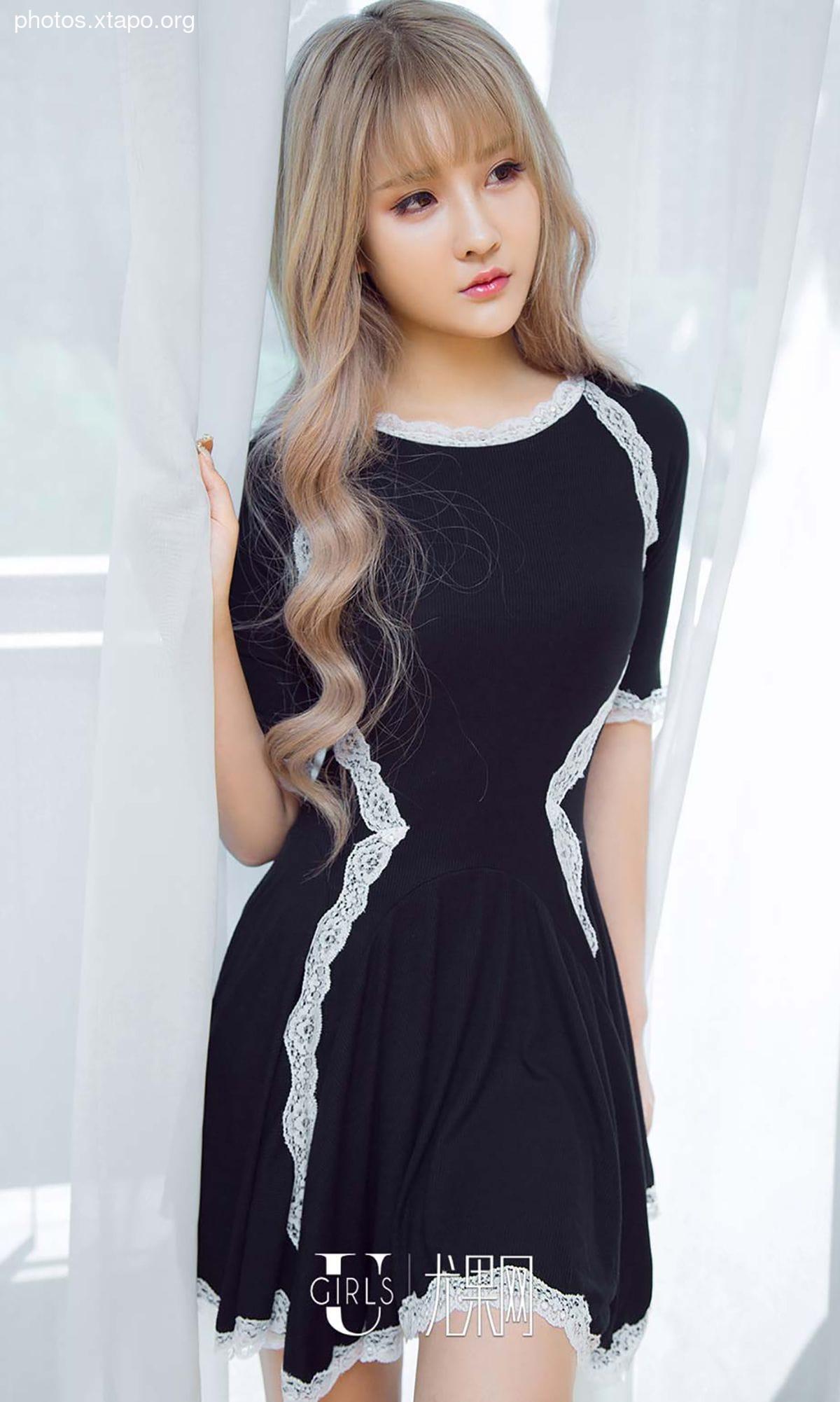 A young woman poses in a stylish black dress adorned with delicate white lace. The dress features a flattering A-line silhouette and short sleeves. The woman's blonde hair is styled in soft waves, framing her face. The backdrop is a simple white curtain, providing a clean and elegant setting.