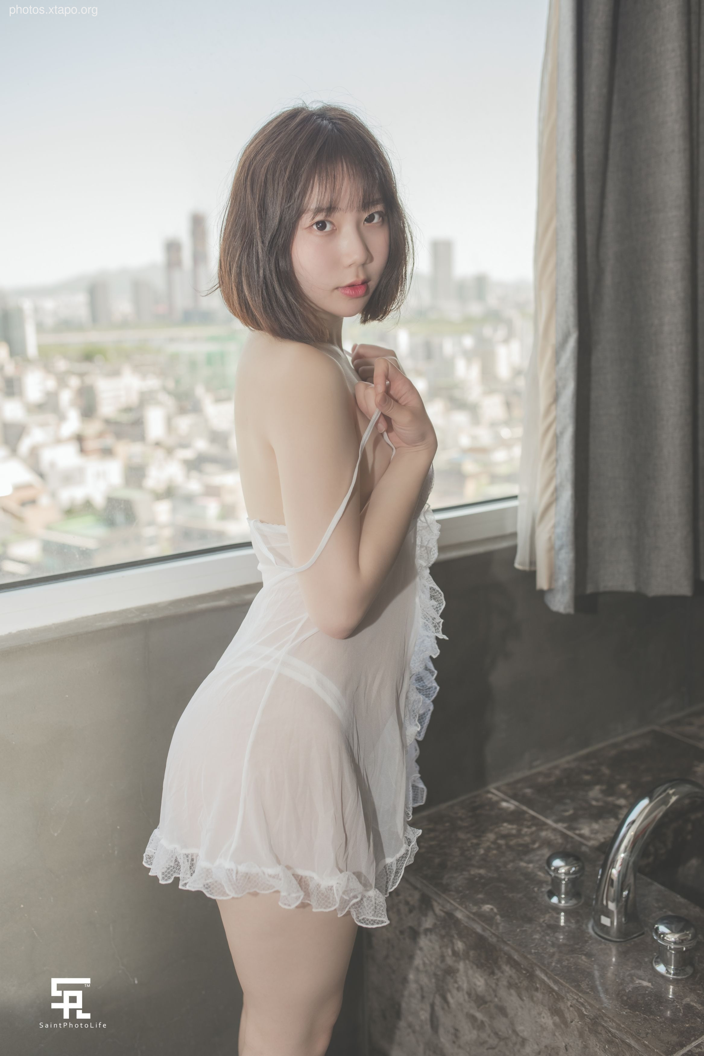 A young woman in a white lace slip dress stands in front of a window with a city view.
