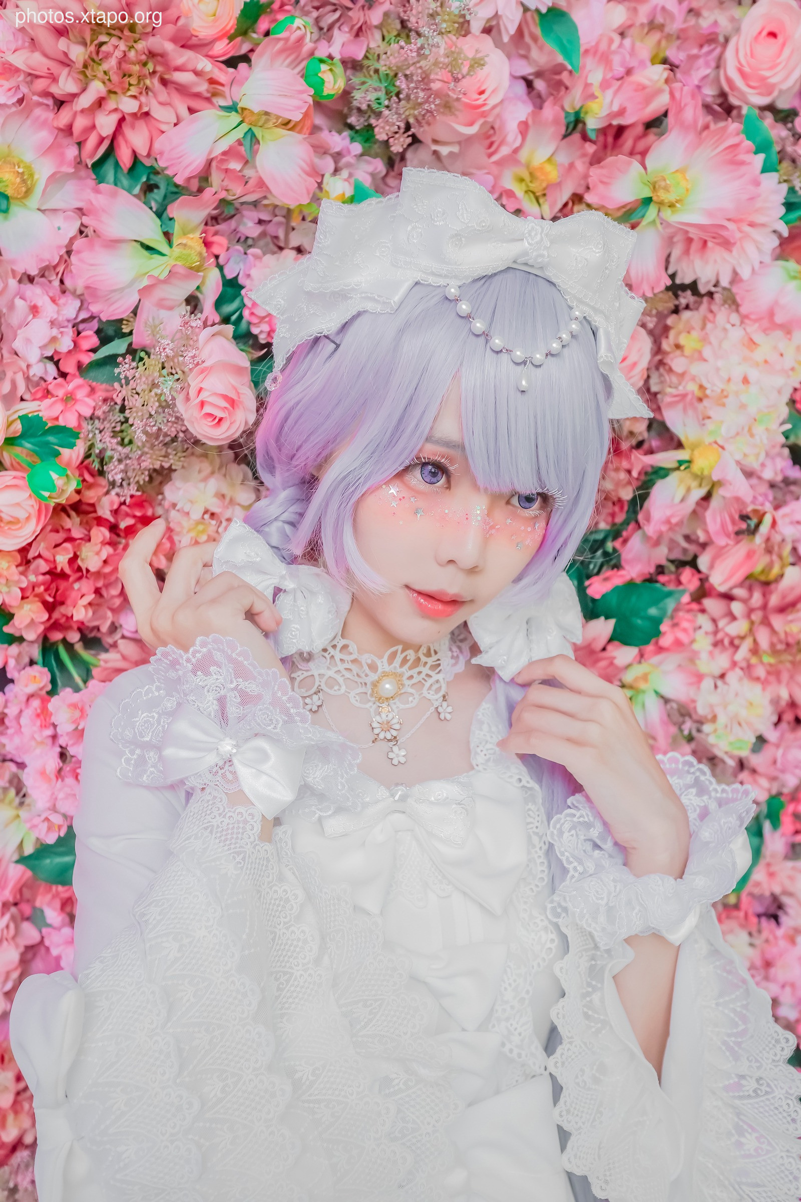 A young woman in a beautiful white lolita dress poses amidst a vibrant backdrop of pink flowers. Her lavender wig and delicate accessories add to her enchanting appearance.