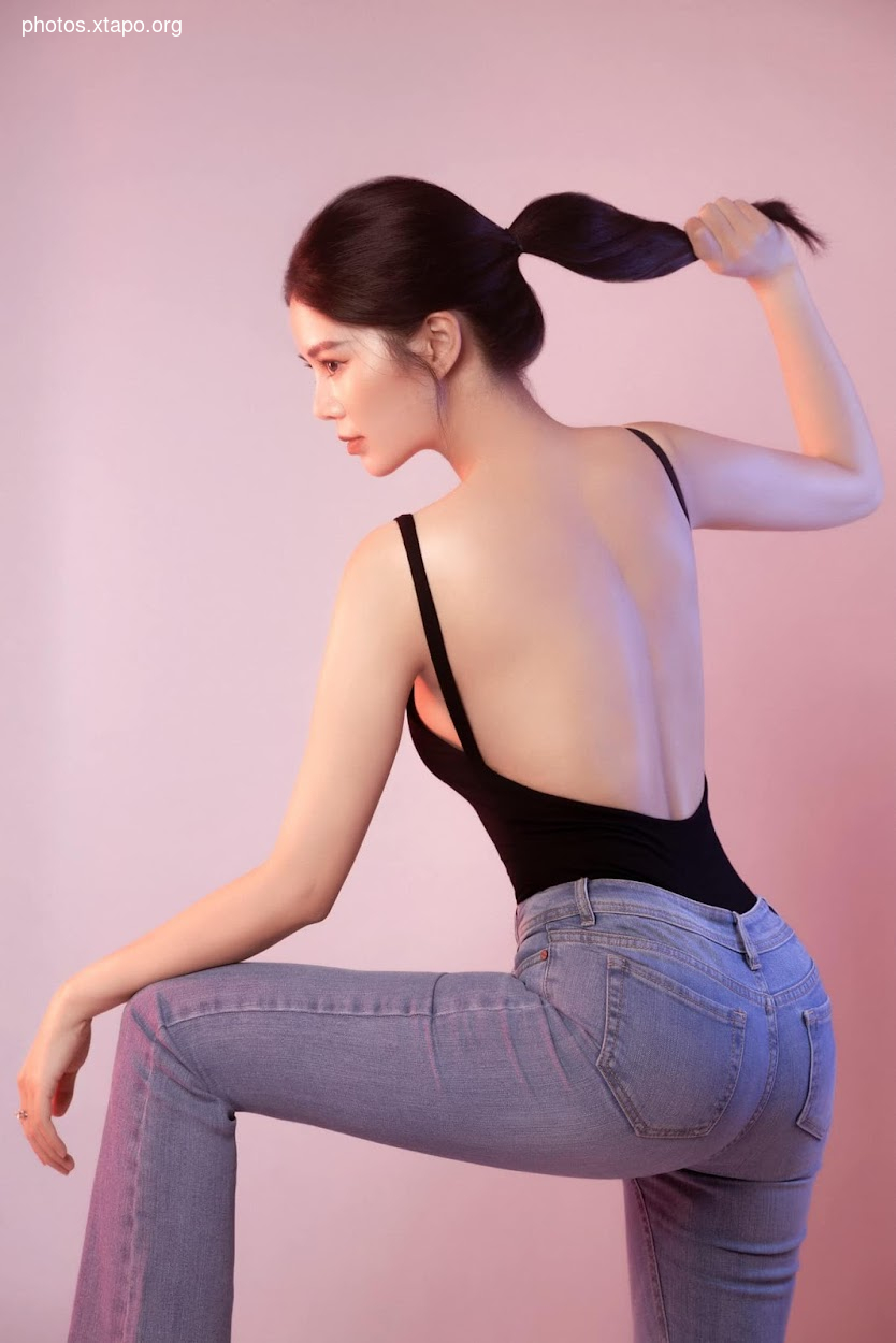 A woman with her hair in a ponytail wears a black bodysuit and light blue jeans. She is posed against a pink background.