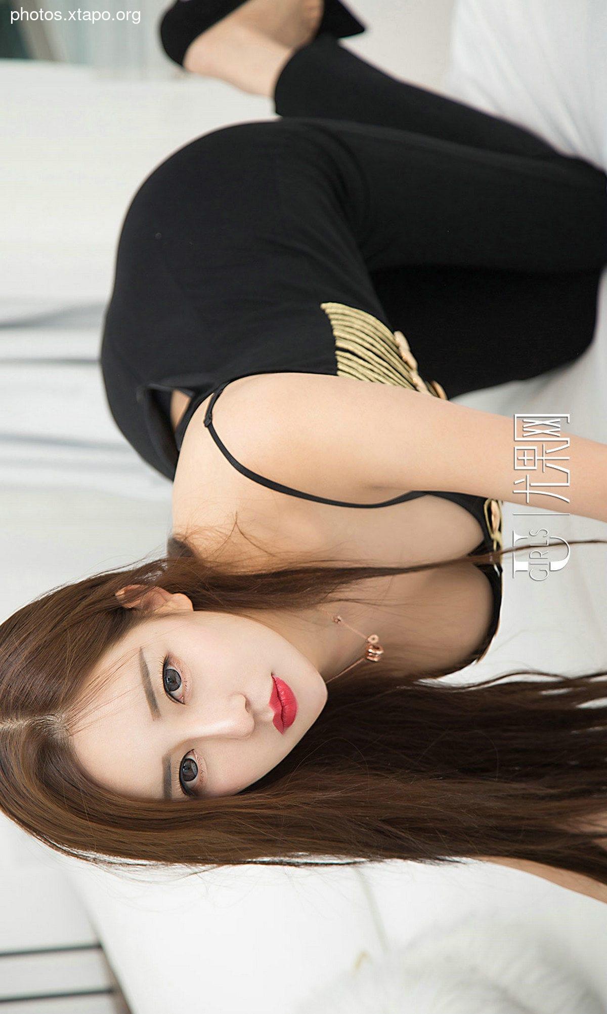 A young woman with long brown hair lies on her stomach wearing a black dress with gold accents.