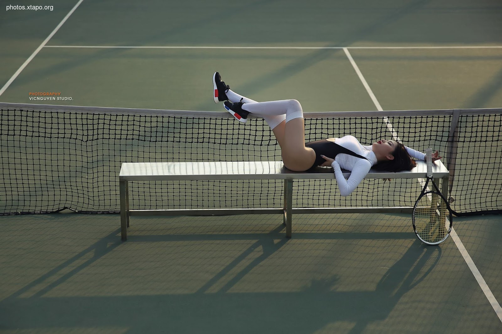 A woman takes a break from her tennis match, relaxing on a bench with her racket nearby.