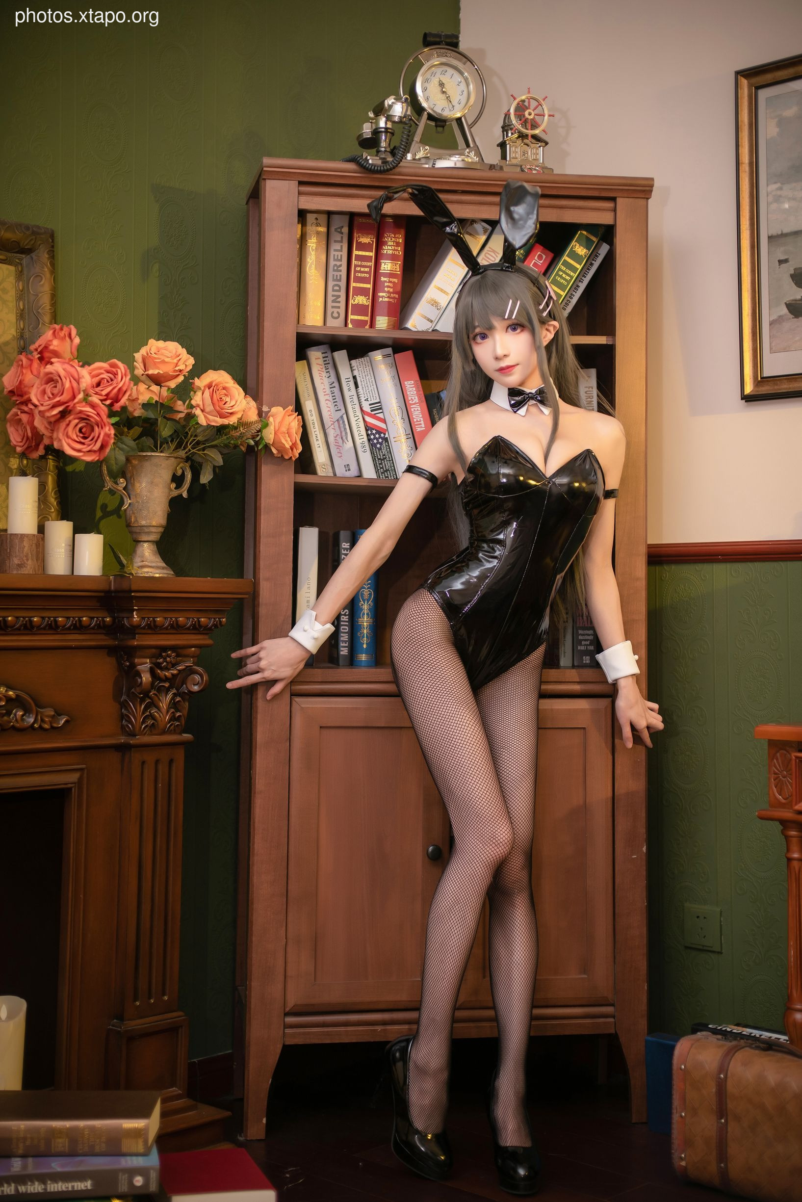 A young woman dressed in a black bunny girl costume poses in a library setting. She is leaning against a wooden bookcase filled with books. The image is suggestive but not explicit.