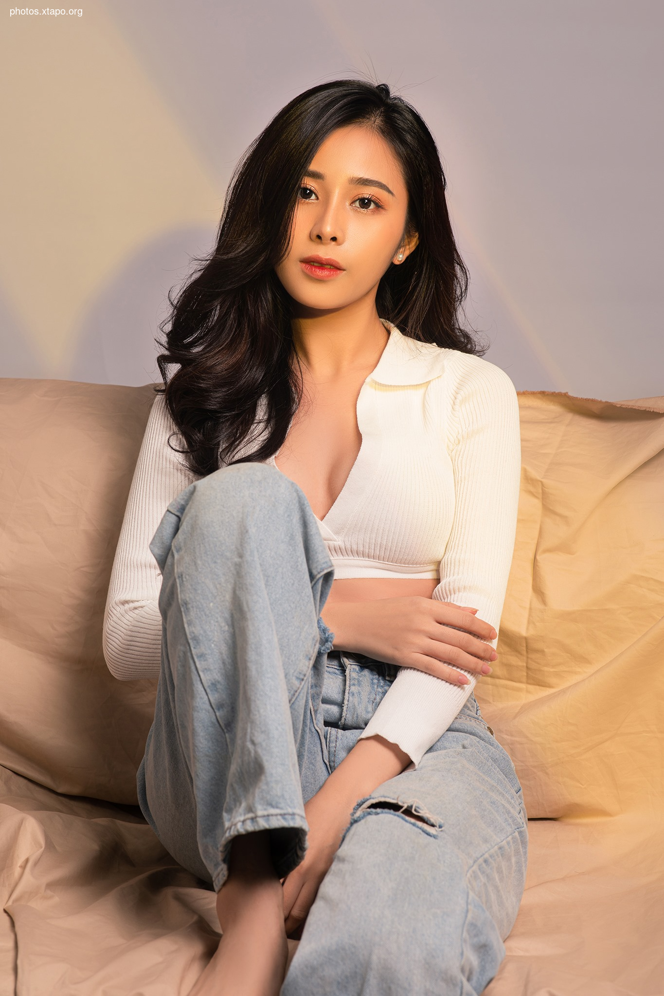 A young woman with long dark hair sits on a beige couch, wearing a white ribbed collared sweater and light blue jeans.  She is looking directly at the camera.
