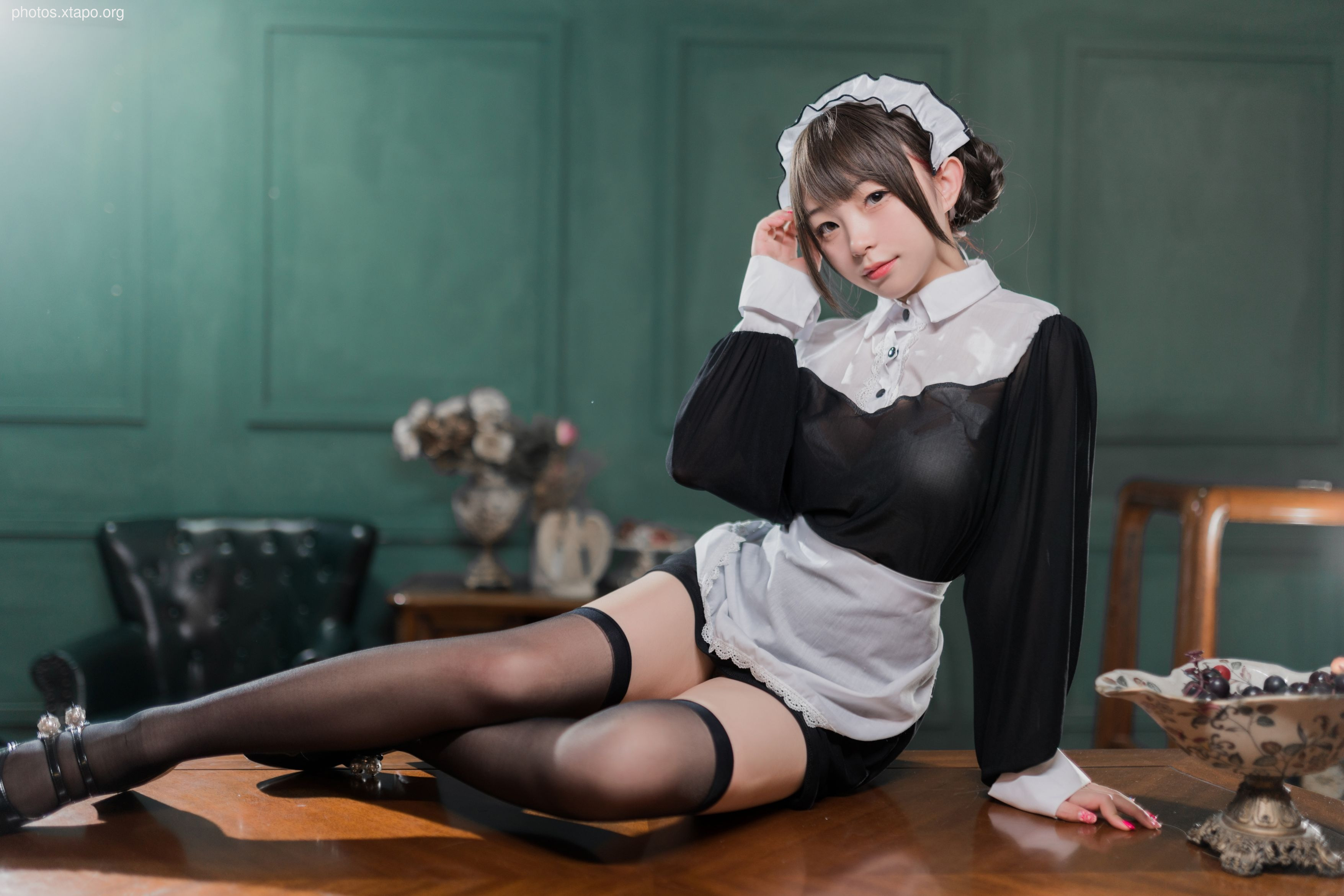 A young woman dressed as a maid poses for a photo. She is wearing a black and white maid outfit and black stockings. She is sitting on a table with a bowl of fruit next to her.