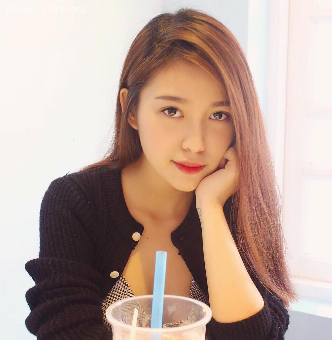 A young woman with long brown hair sits at a table, enjoying a cup of boba tea. She is wearing a black cardigan and a patterned top.